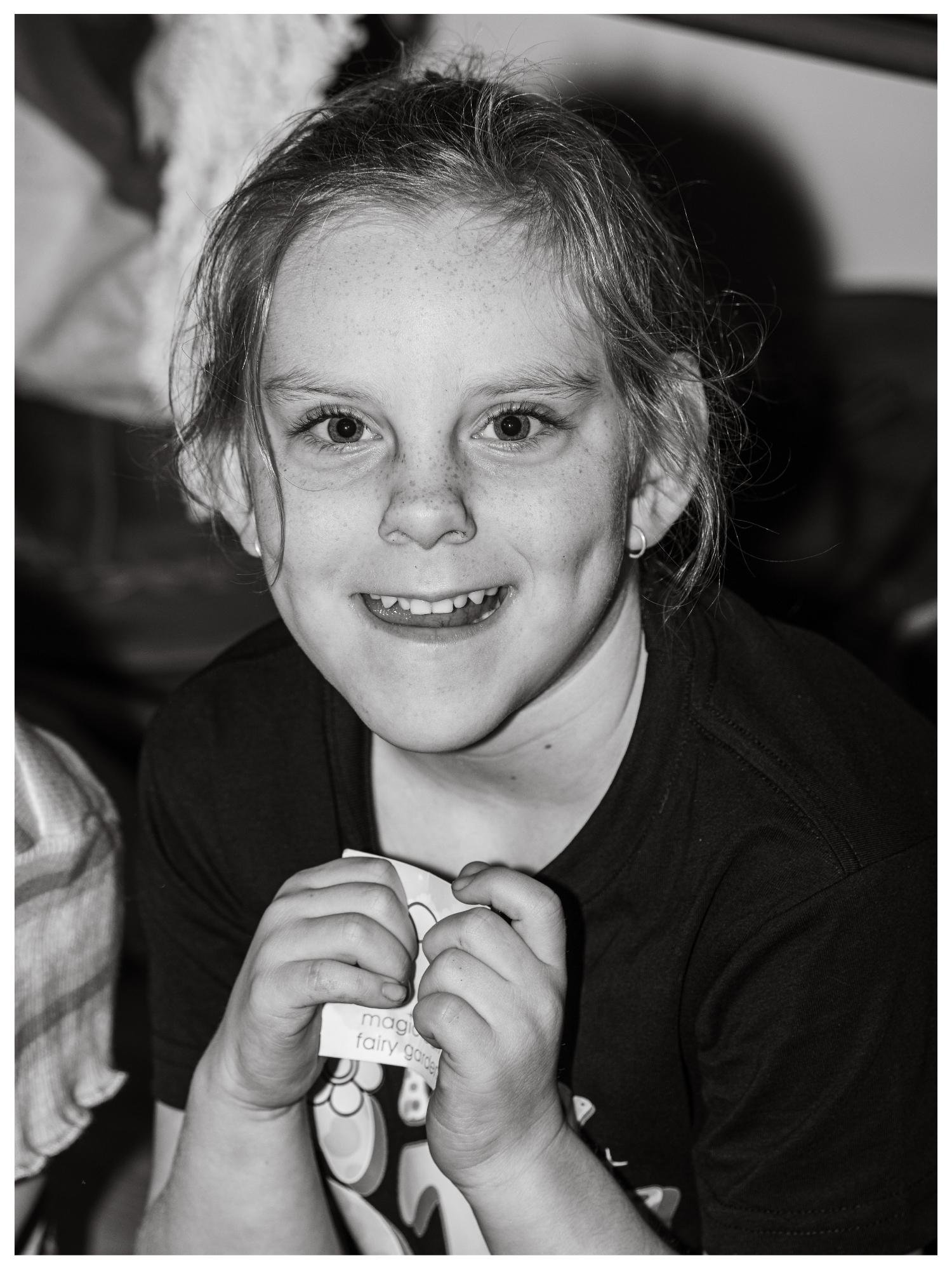 A smiling young girl with light in her eyes, holding a small card with the words 'magical fairy garden' printed on it.