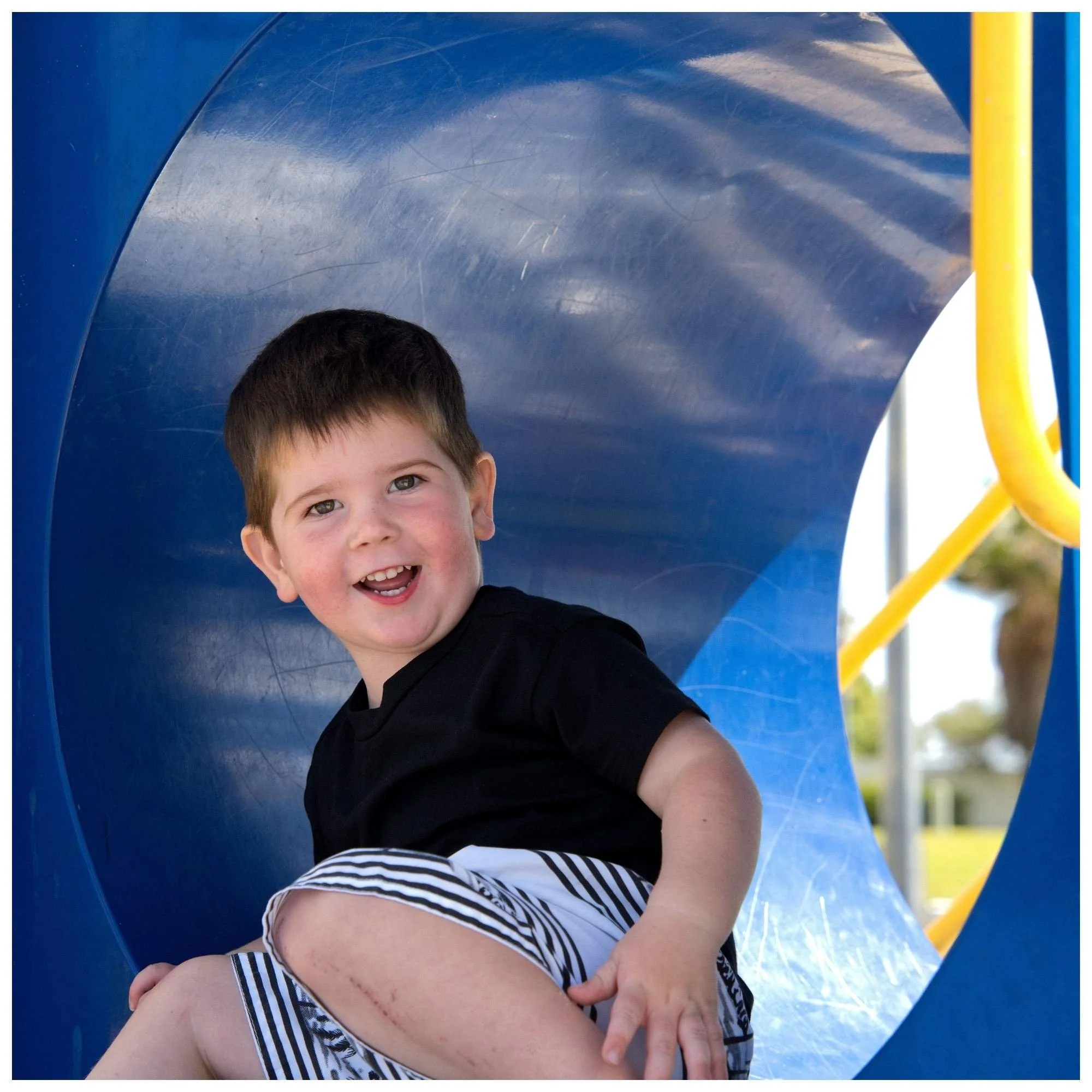 Young boy with brown hair and a big smile sitting inside a blue playground tunnel, wearing a black t-shirt and striped shorts.