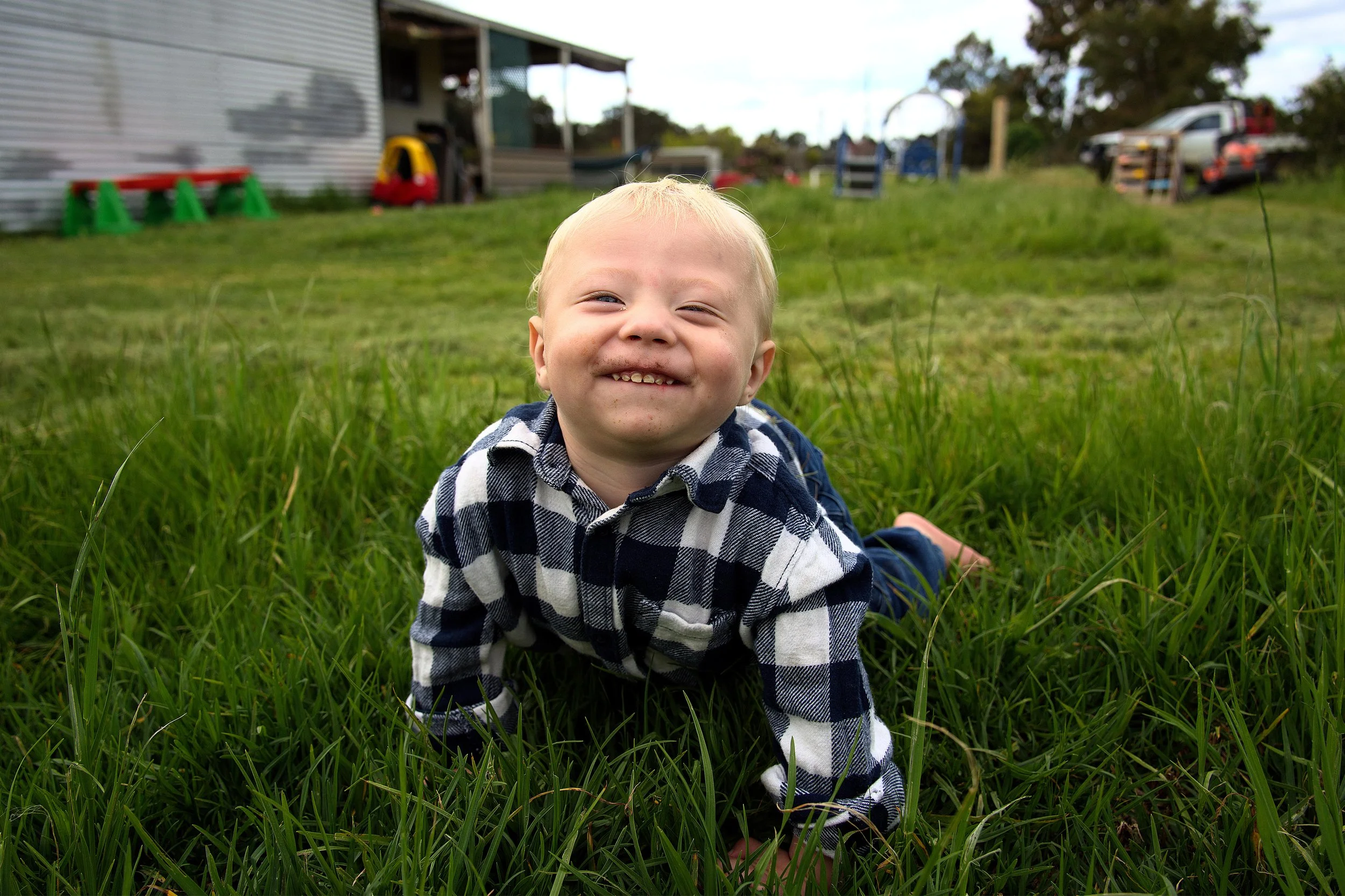 A smiling young boy with blonde hair crawling on green grass, wearing a black and white checkered shirt.
