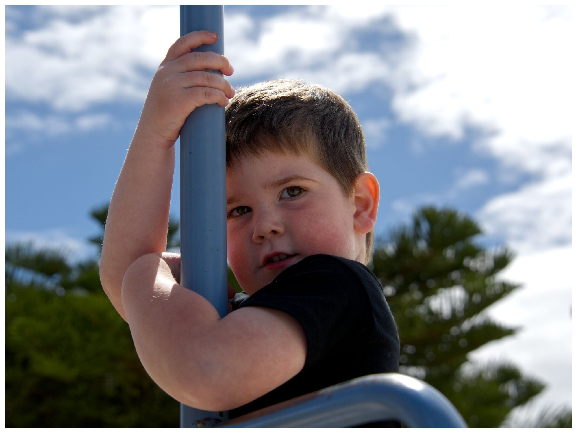 Young boy with short brown hair and fair skin looking over a metal railing outdoors against a backdrop of blue sky, clouds, and green trees.