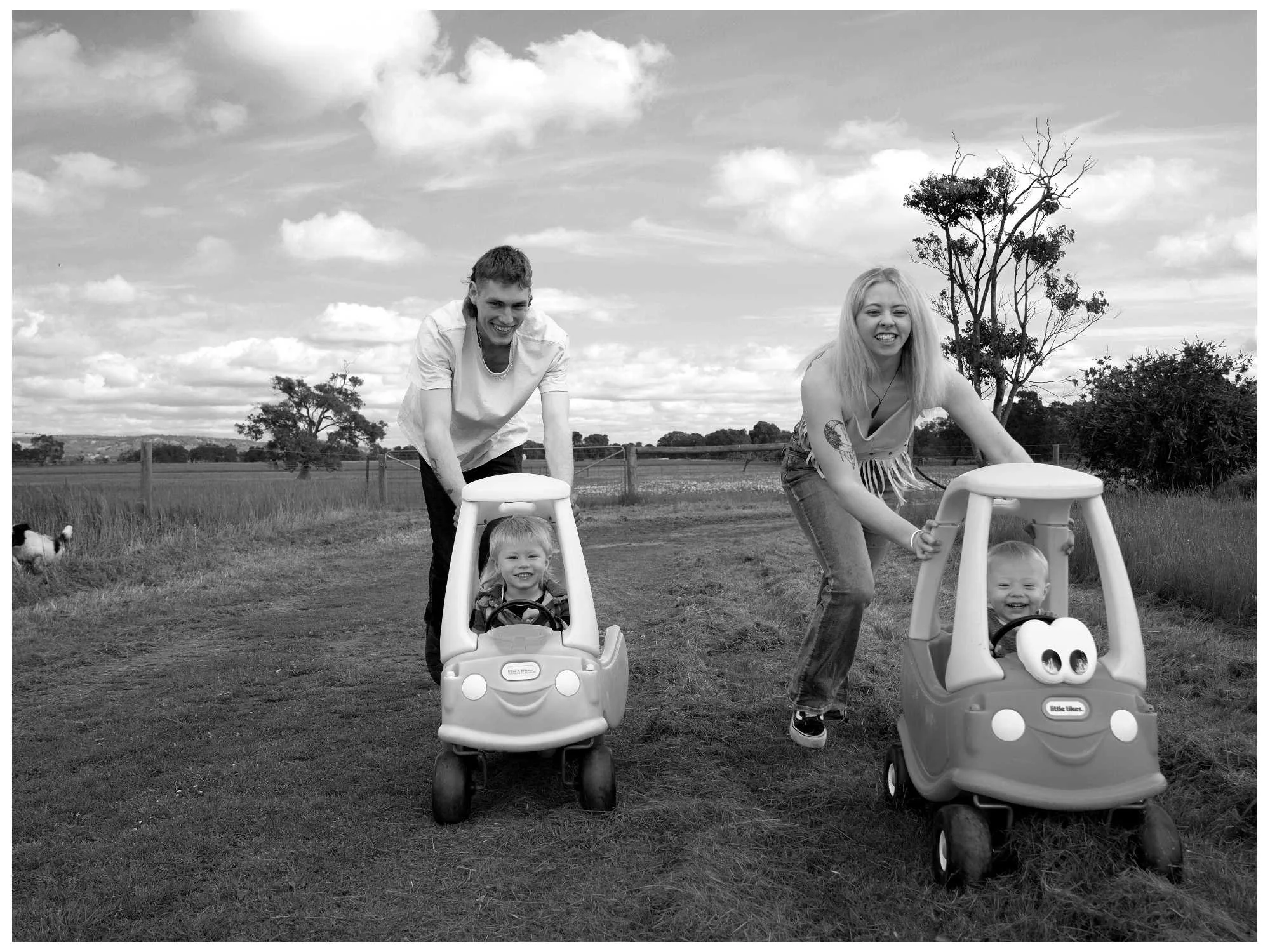 Two people, a man and a woman, are pushing children in toy cars on a grassy field outdoors. The children, a girl and a boy, are smiling and sitting in their toy cars. There are trees and a partly cloudy sky in the background.