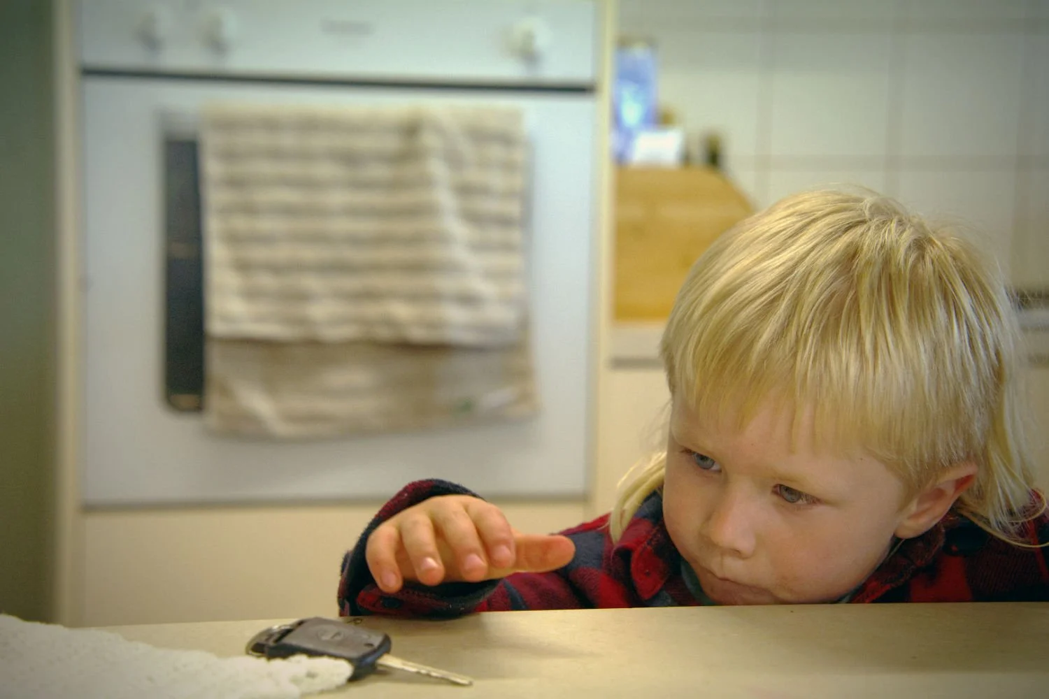 A young boy with blond hair, sitting at a table, looking at a car key on the table with a thoughtful expression in a kitchen