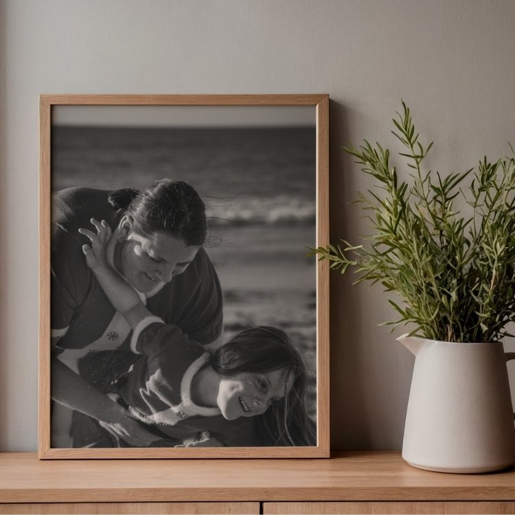 A framed black and white photograph of a woman and a young girl playing on the beach, leaning toward each other and smiling, next to a potted plant on a wooden surface.