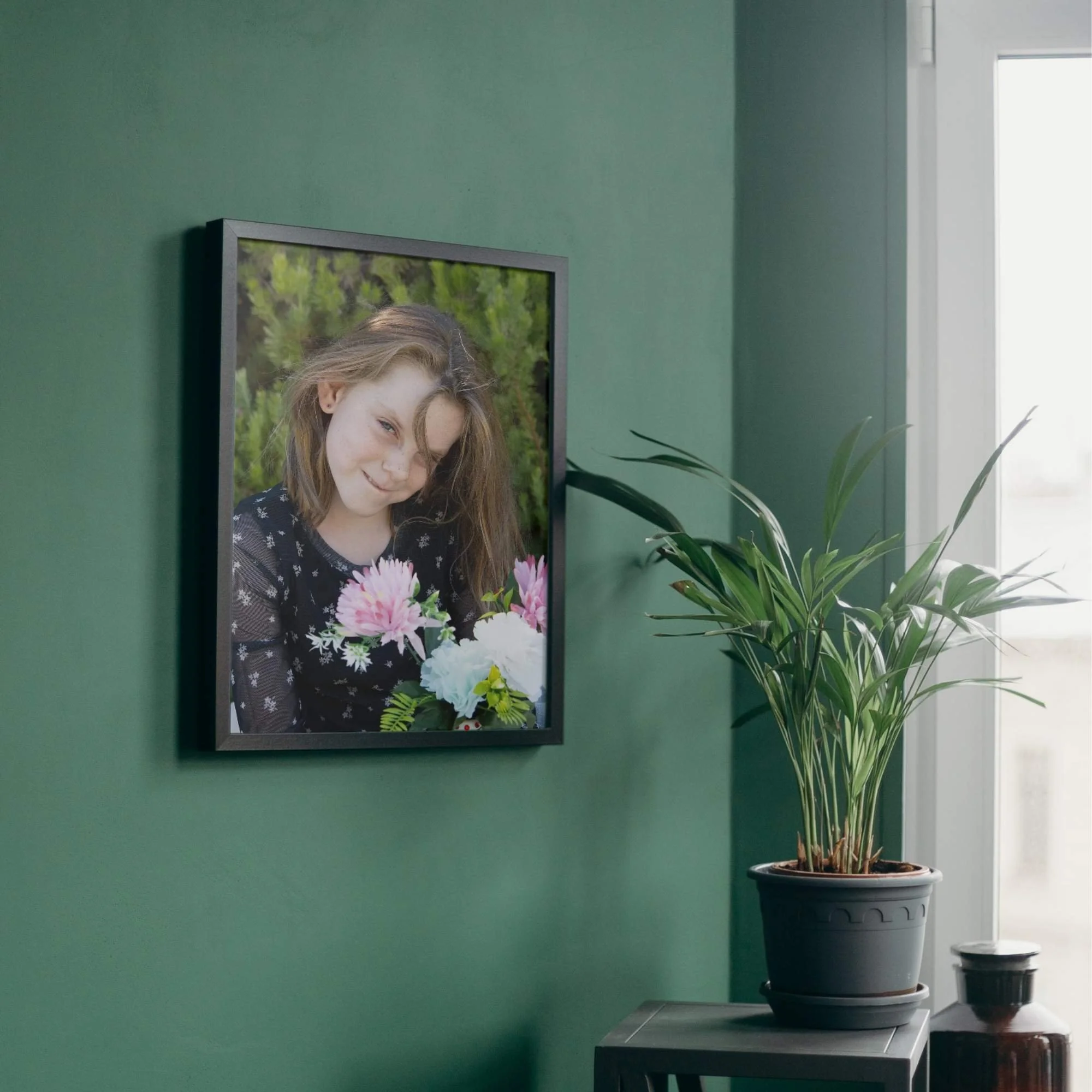 A photograph of a girl holding flowers and smiling is framed and hung on a deep green wall. Nearby a palm plant sits in a pot by the window.