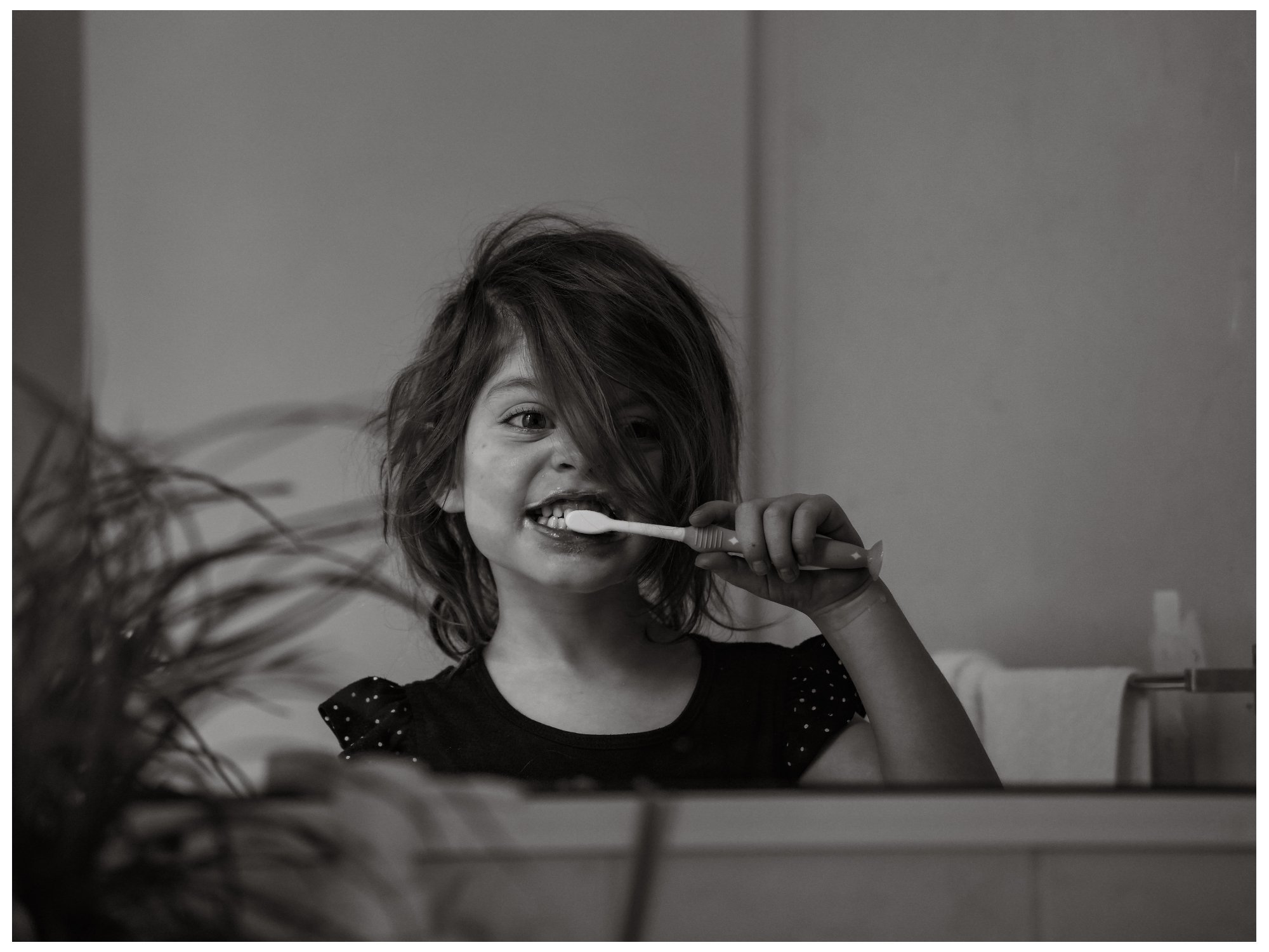 A young girl brushing her teeth in front of a mirror with a big smile and hair partially covering her face.