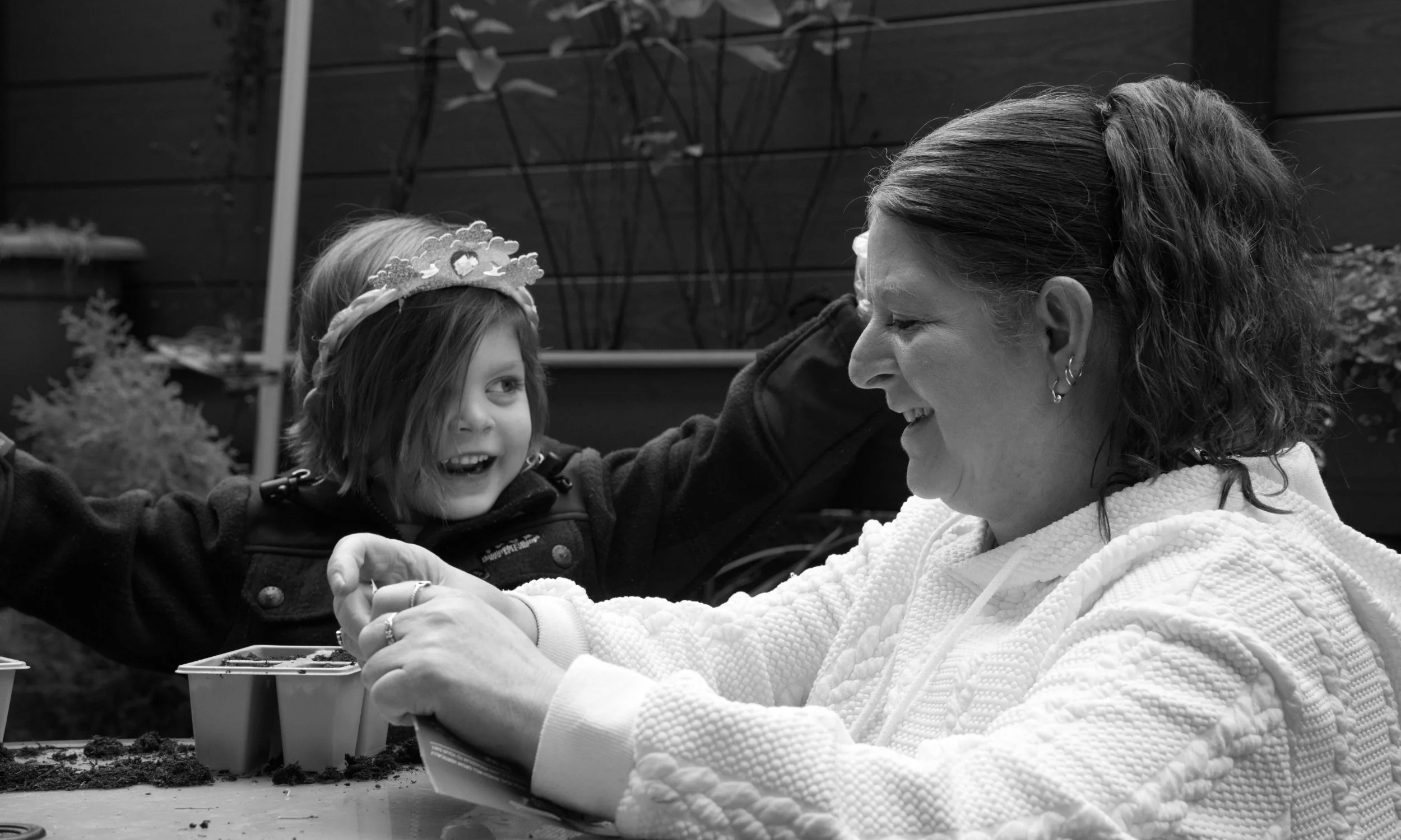 A woman and a young girl with a crown around her head having fun planting flowers together at an outdoor table in a home documentary family photography session.