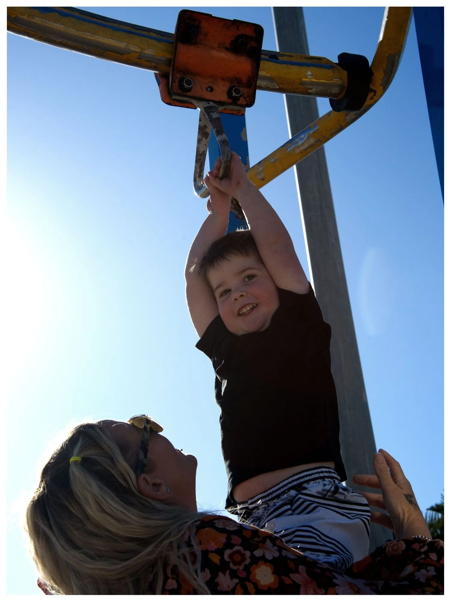 A young boy hanging from a playground bar with an adult woman supporting him, under clear blue sky.