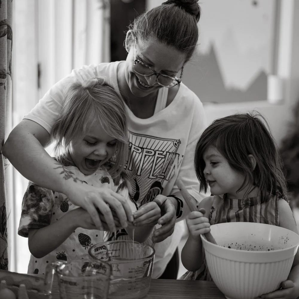 A woman and two young girls smiling and laughing while cooking or baking together in a kitchen, with one girl cracking eggs into a measuring cup and the other girl stirring a bowl.