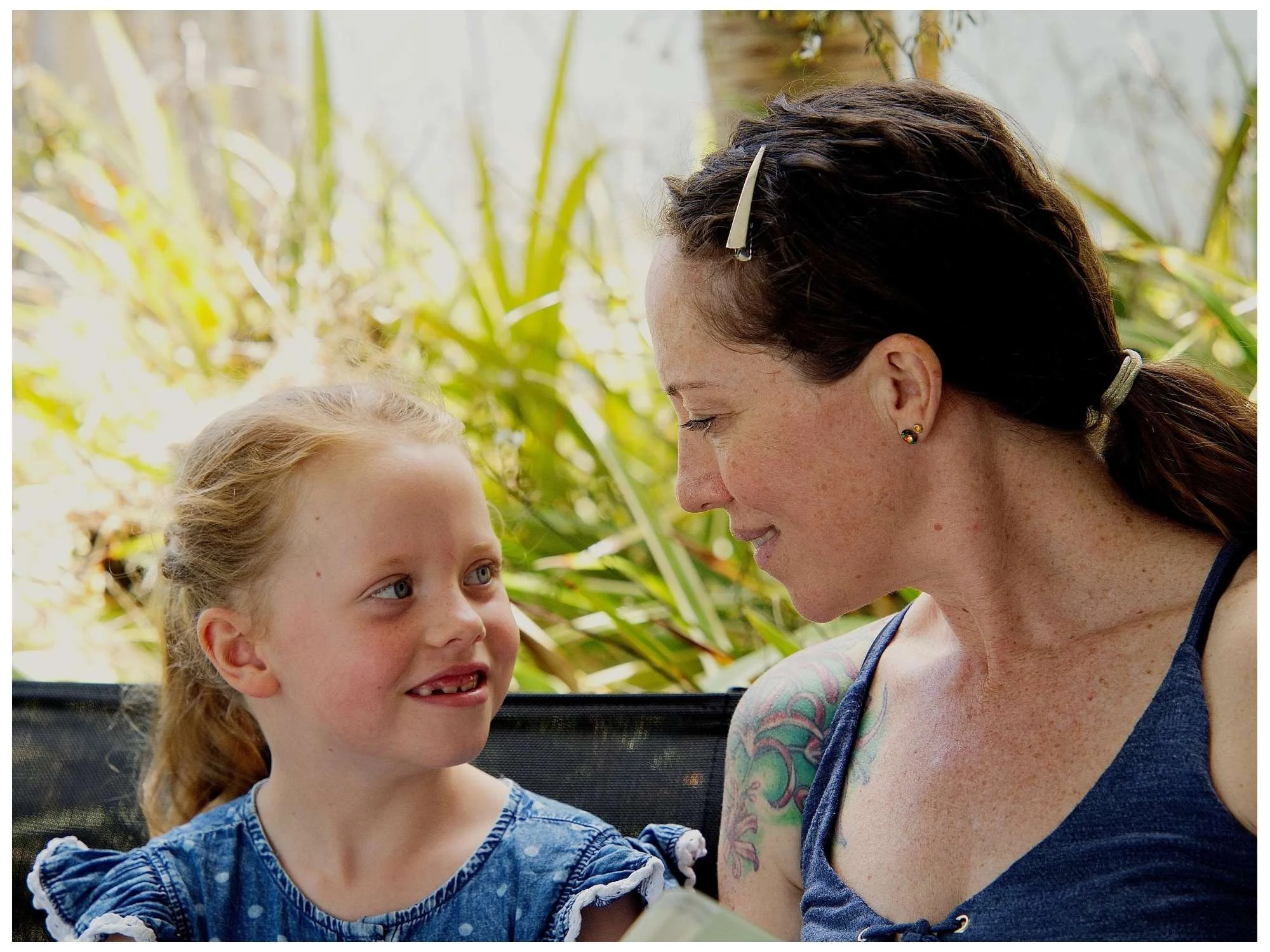 A woman and a young girl are sitting close together outdoors, smiling and looking at each other. The woman has dark hair, freckles, and tattoos on her arm. The girl has red hair and is wearing a blue dress, and they appear to be sharing a happy momen
