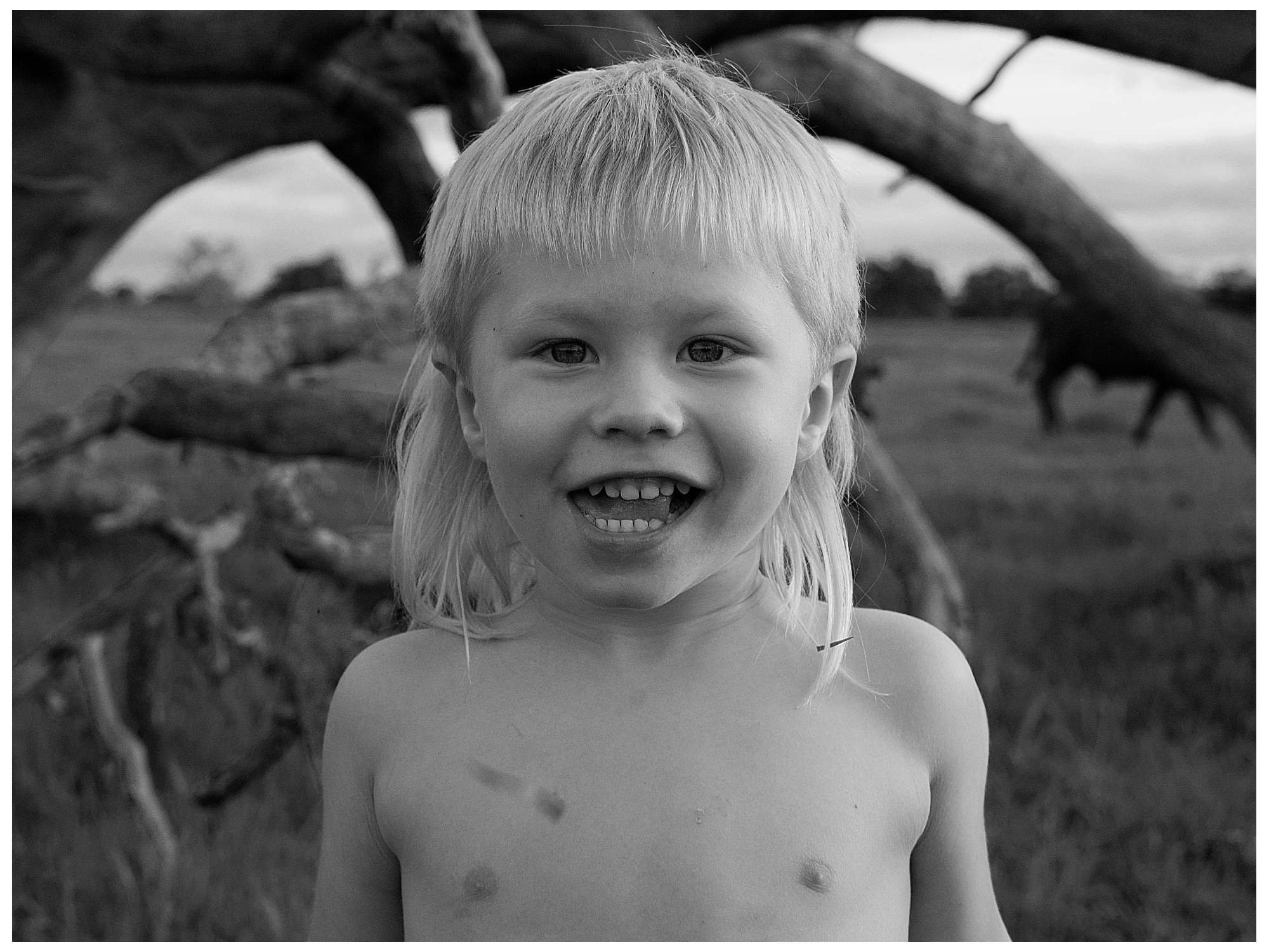 A young boy with shoulder-length light hair smiling and showing teeth, shirtless, standing outdoors in front of a large, fallen tree in a grassy field.