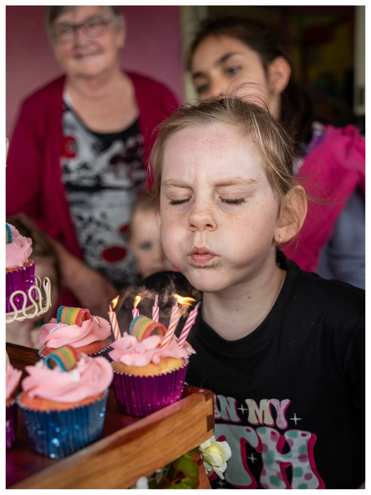 A girl with light skin, freckles, and closed eyes is blowing out candles on pink frosted cupcakes. Two women and a child are in the background, watching the celebration.