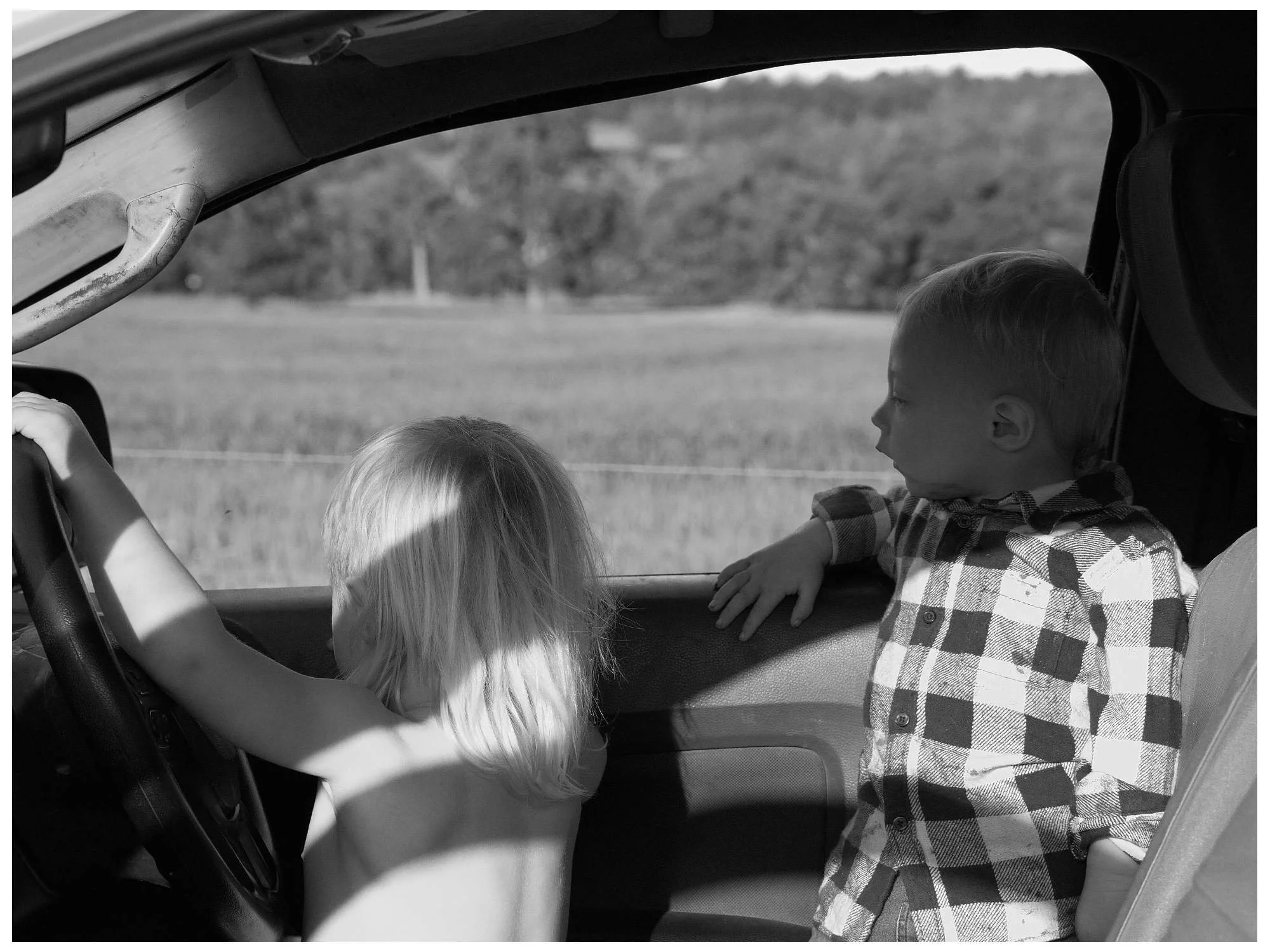 Two young children inside a vehicle, one boy with blonde hair is the driver, and a boy with short hair and a checkered shirt leans on the window, looking outside at a countryside scene.