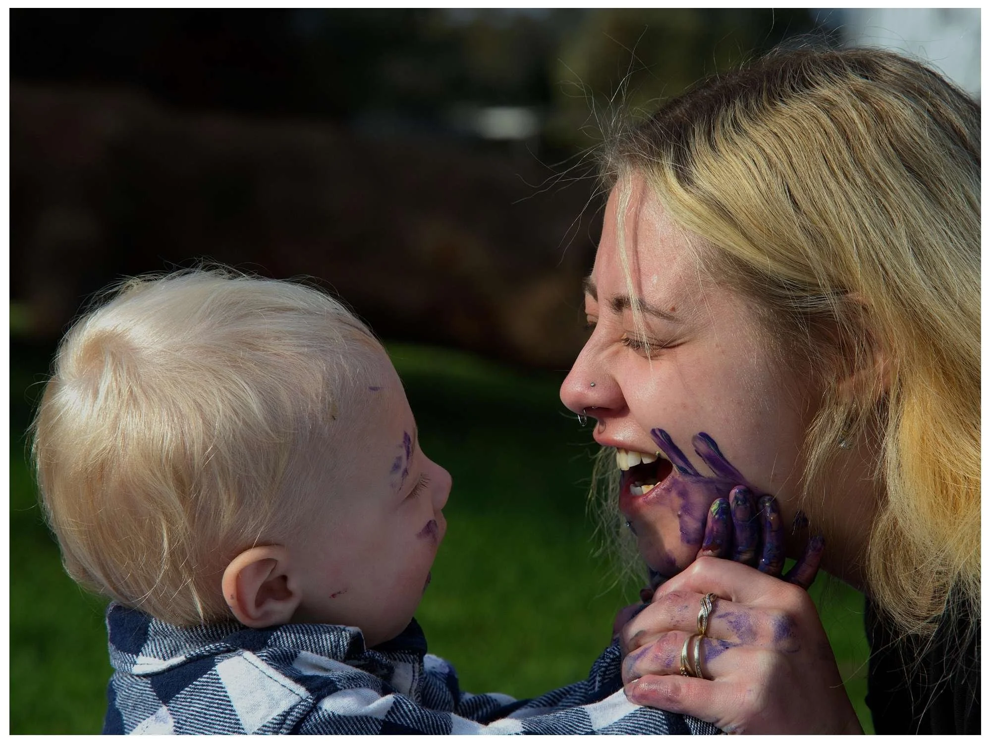 A woman and a young child with paint on their faces and hands, smiling and playing together outside.