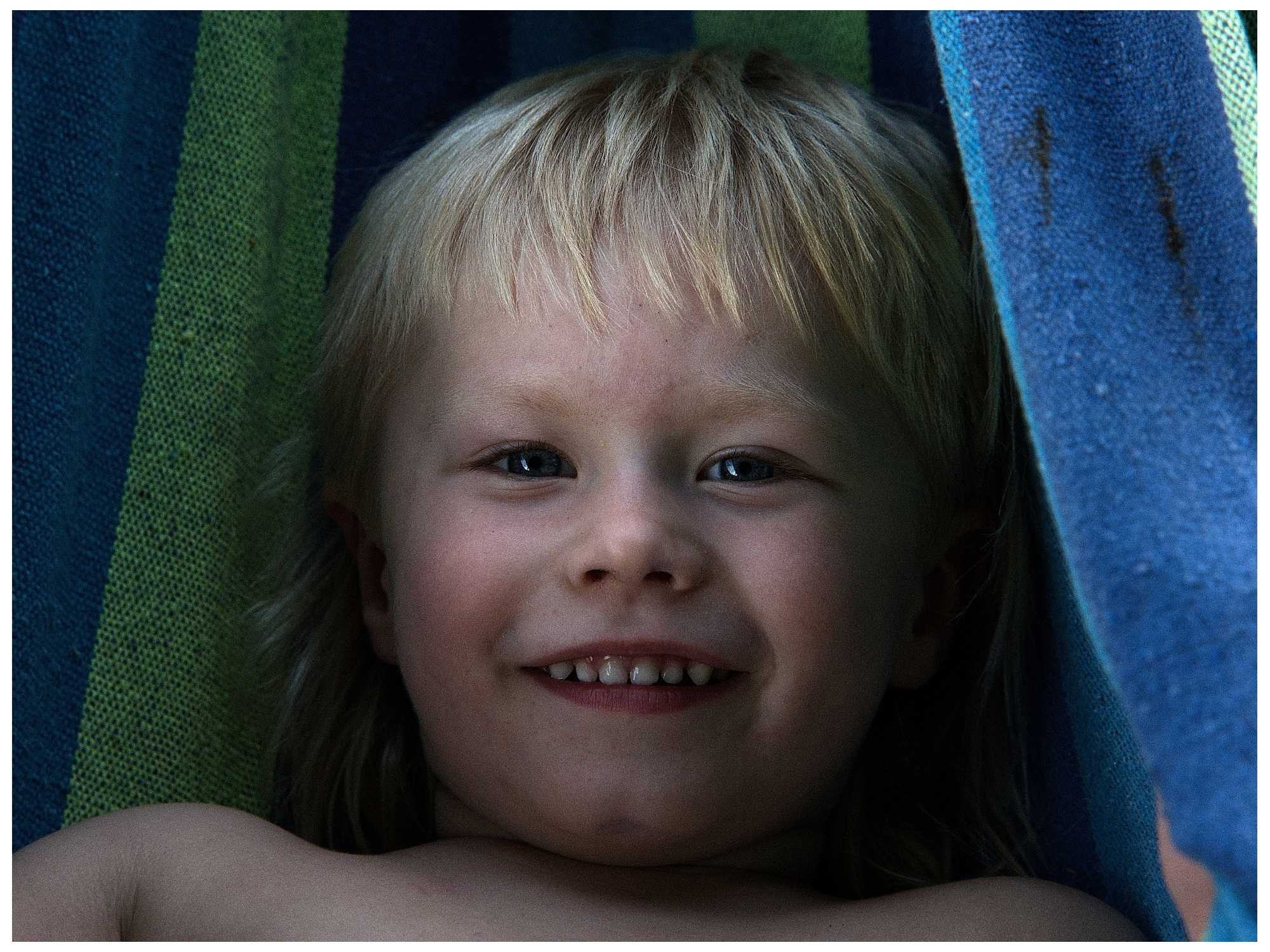 A young boy with blonde hair and blue eyes smiling, lying on a colorful striped towel.