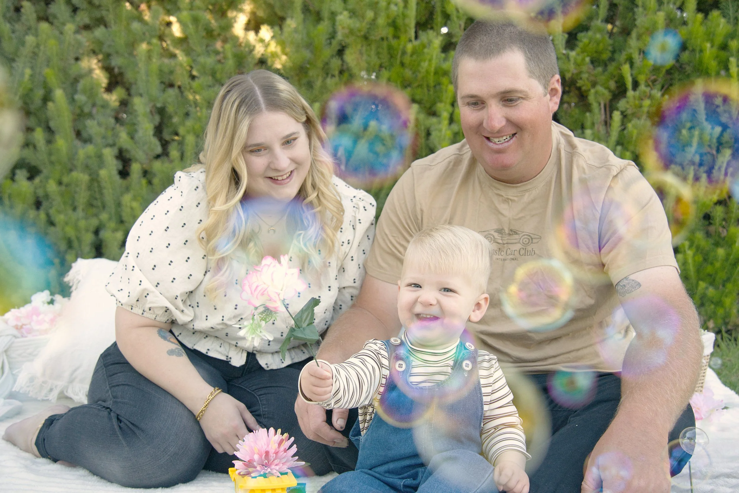 A family of three smile together surrounded by bubbles. The young boy in front holds out a flower