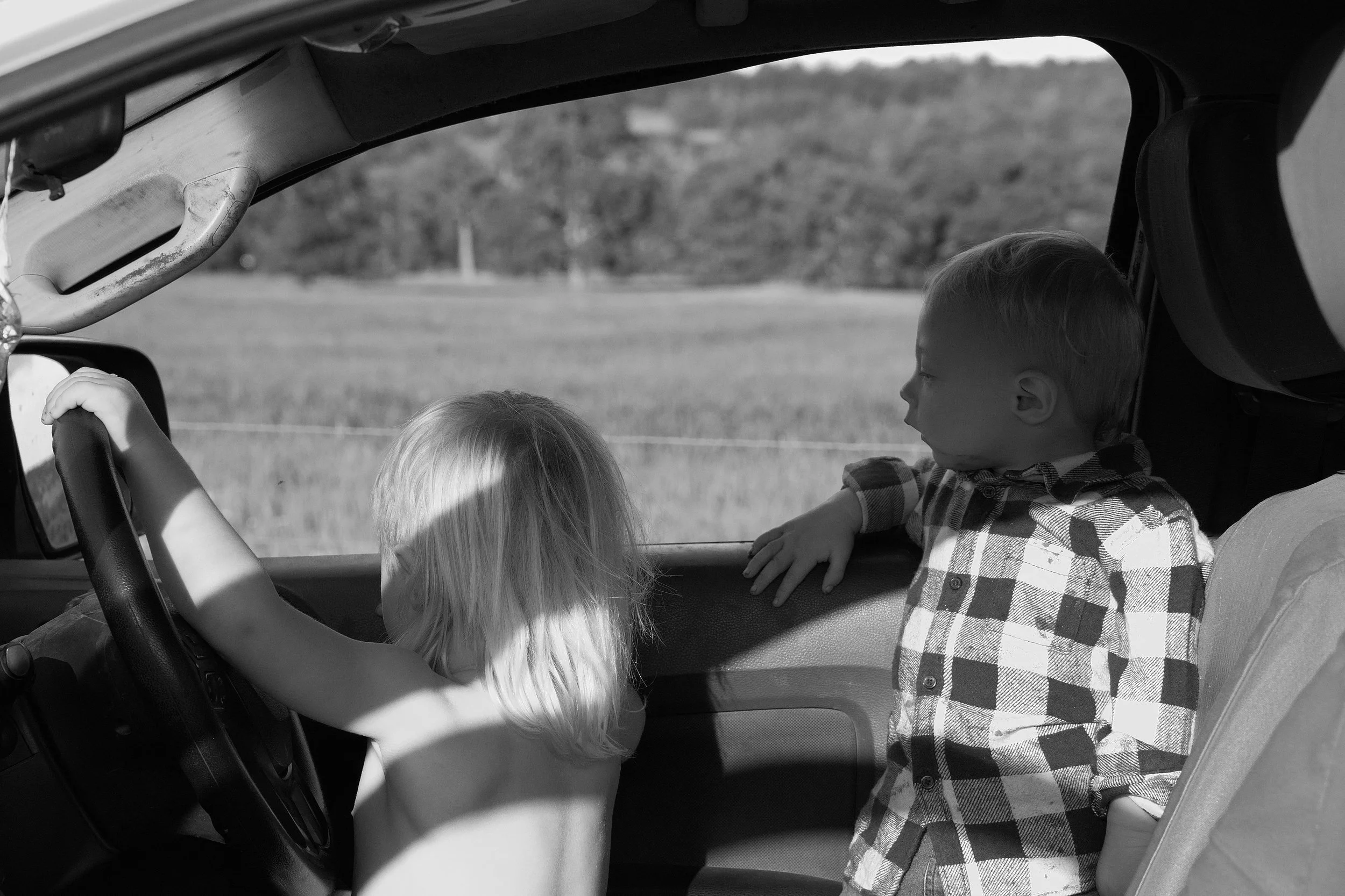 Two young children in a vehicle, one girl with blonde hair on the driver's side and a boy with short hair wearing a plaid shirt on the passenger side, looking at each other while sitting in the front seats of the car.