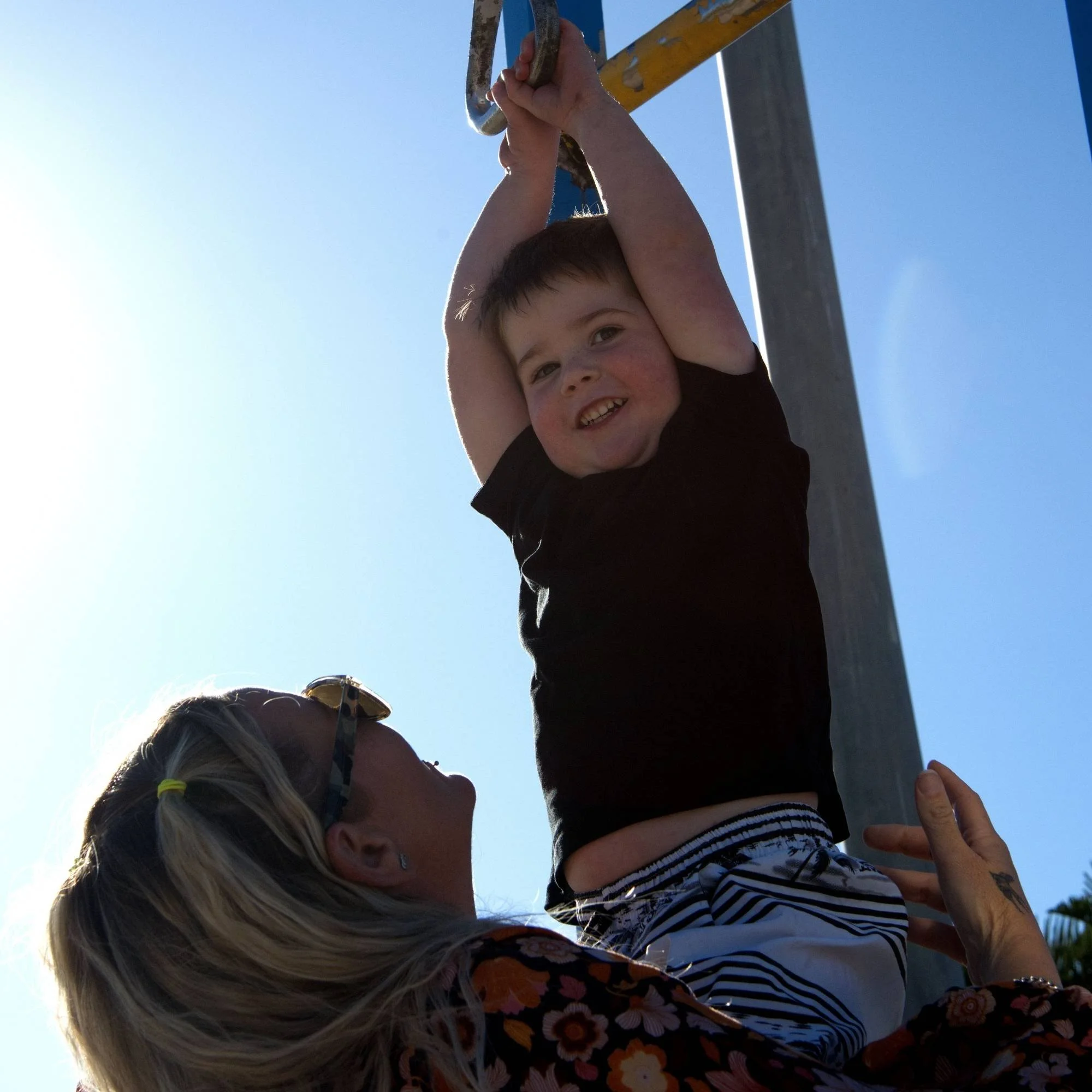 A photo of a boy smiling as his mother lifts him on a flying fox at the park