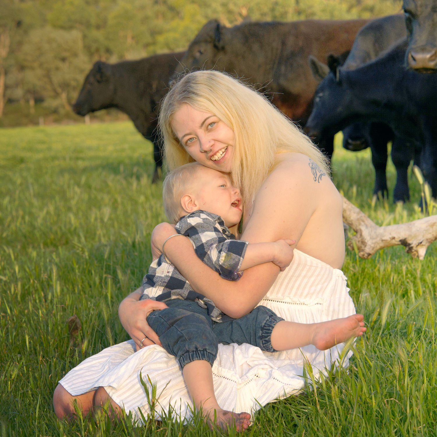 A mother in a white dress sits in a field with her young son. Cows are seen in the background
