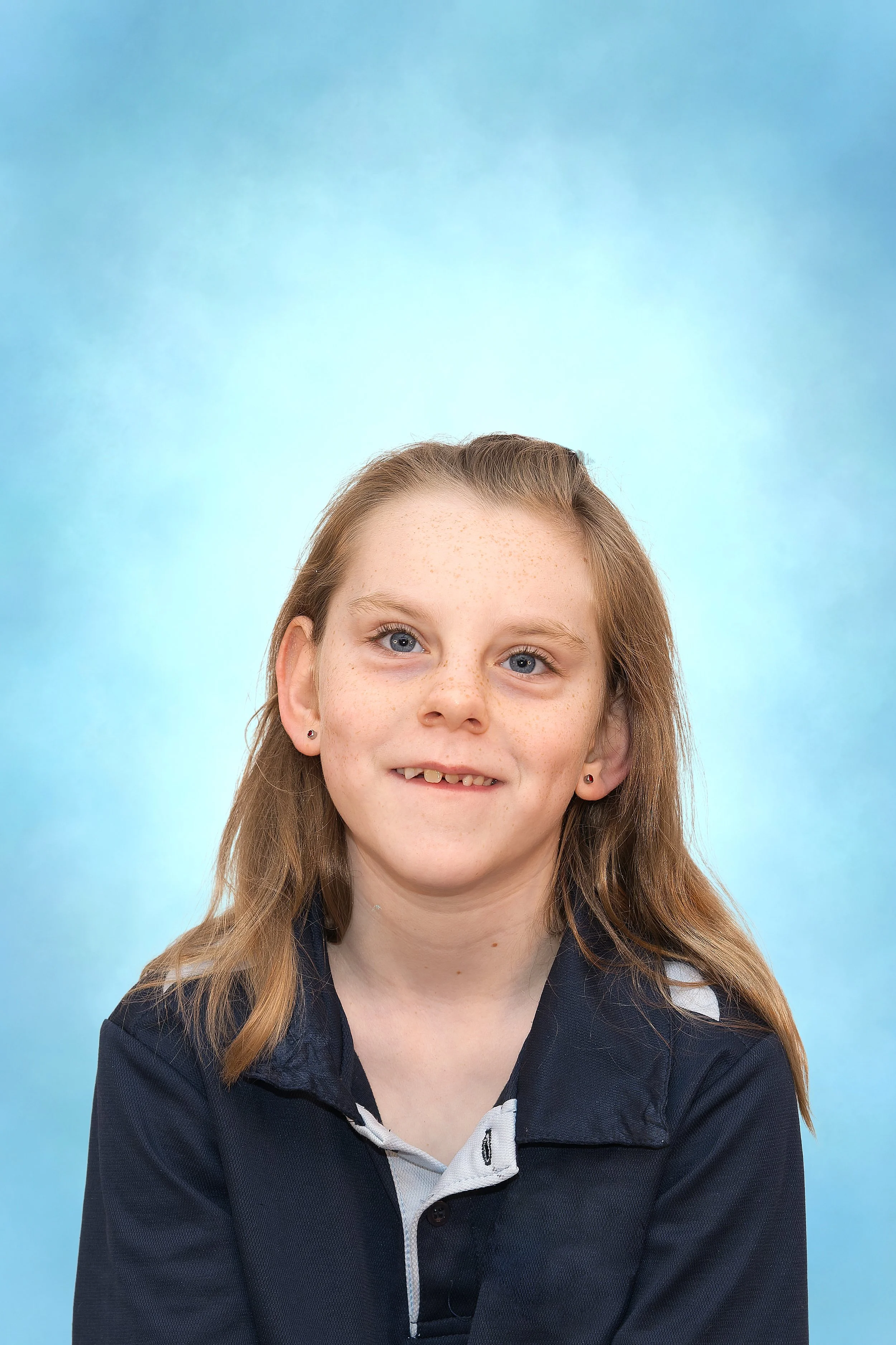 A school-style portrait of a young girl with brown hair in a navy blue polo shirt smilig for the camera in front of a blue background.