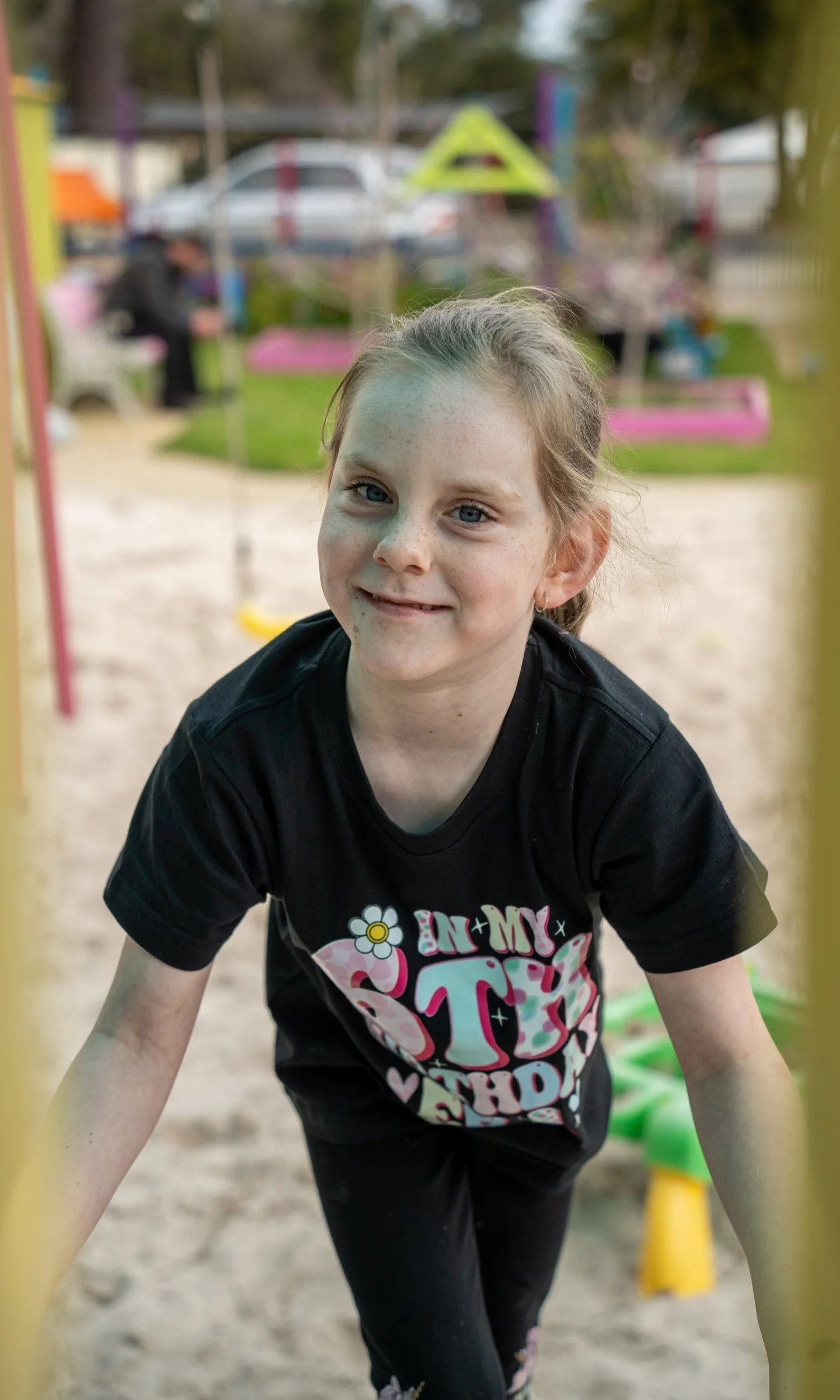 A young girl with light skin, blue eyes, and freckles smiling at the camera, leaning on playground equipment at a park. She is wearing a black T-shirt with colorful text and graphics.