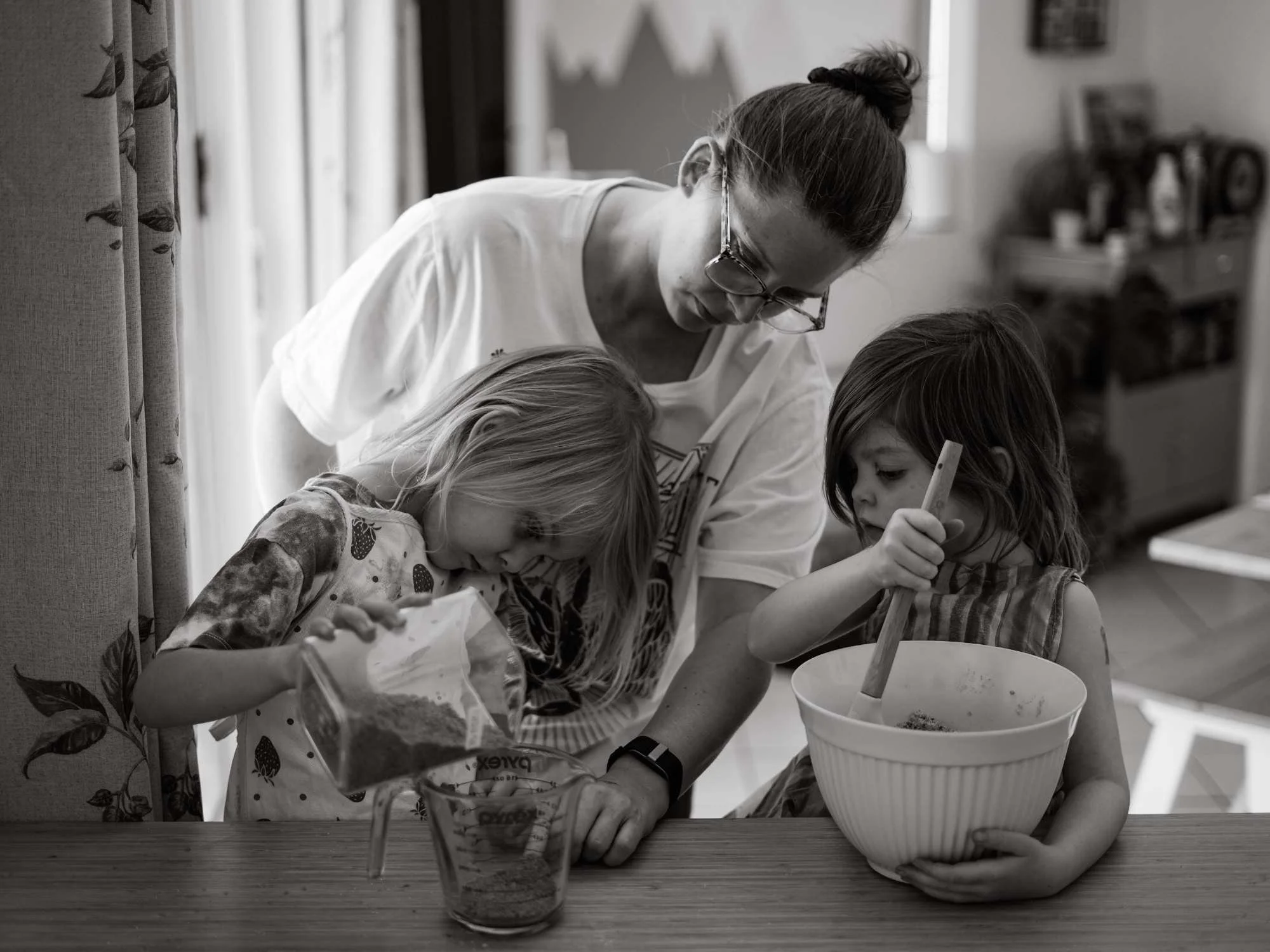 A mum helps two girls baking. One girl mixes in a bowl whilst the other measures ingredients