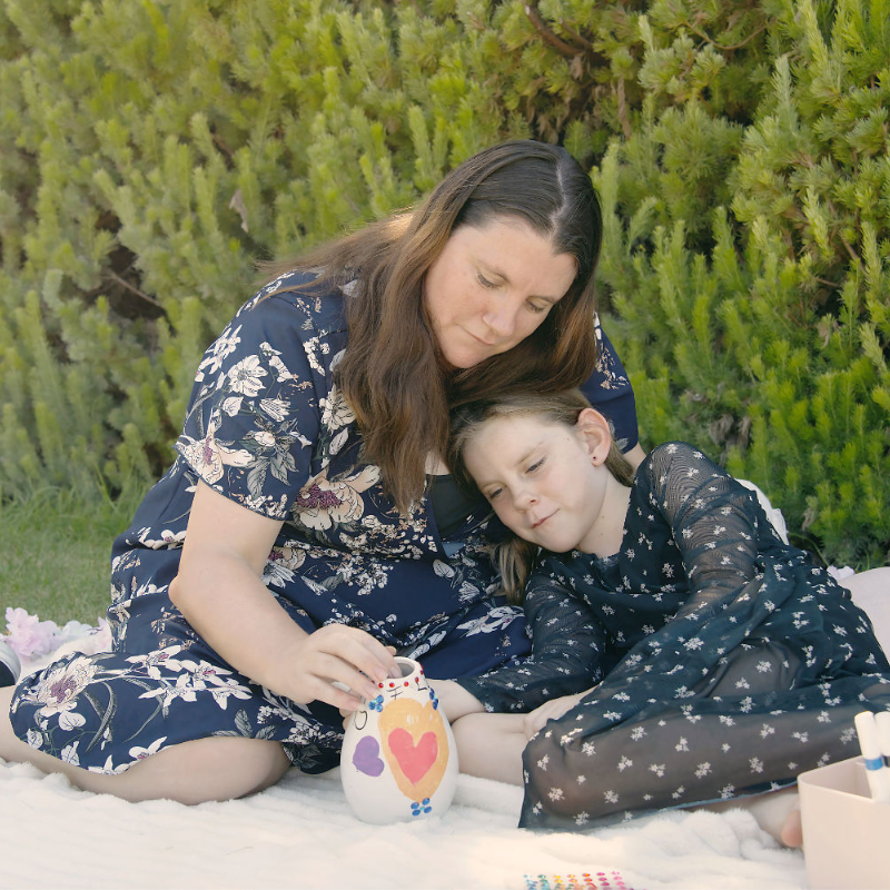 A mother and daughter in navy blue dresses cuddle whilst painting a small vase