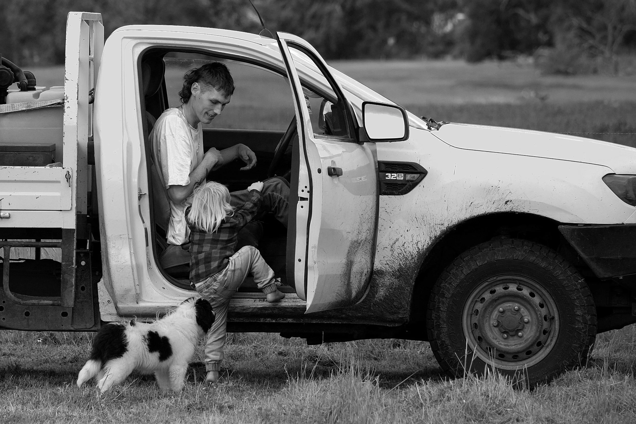 A boy and a girl playing near and inside a pickup truck with muddy sides, with a dog standing nearby, on a grassy field.