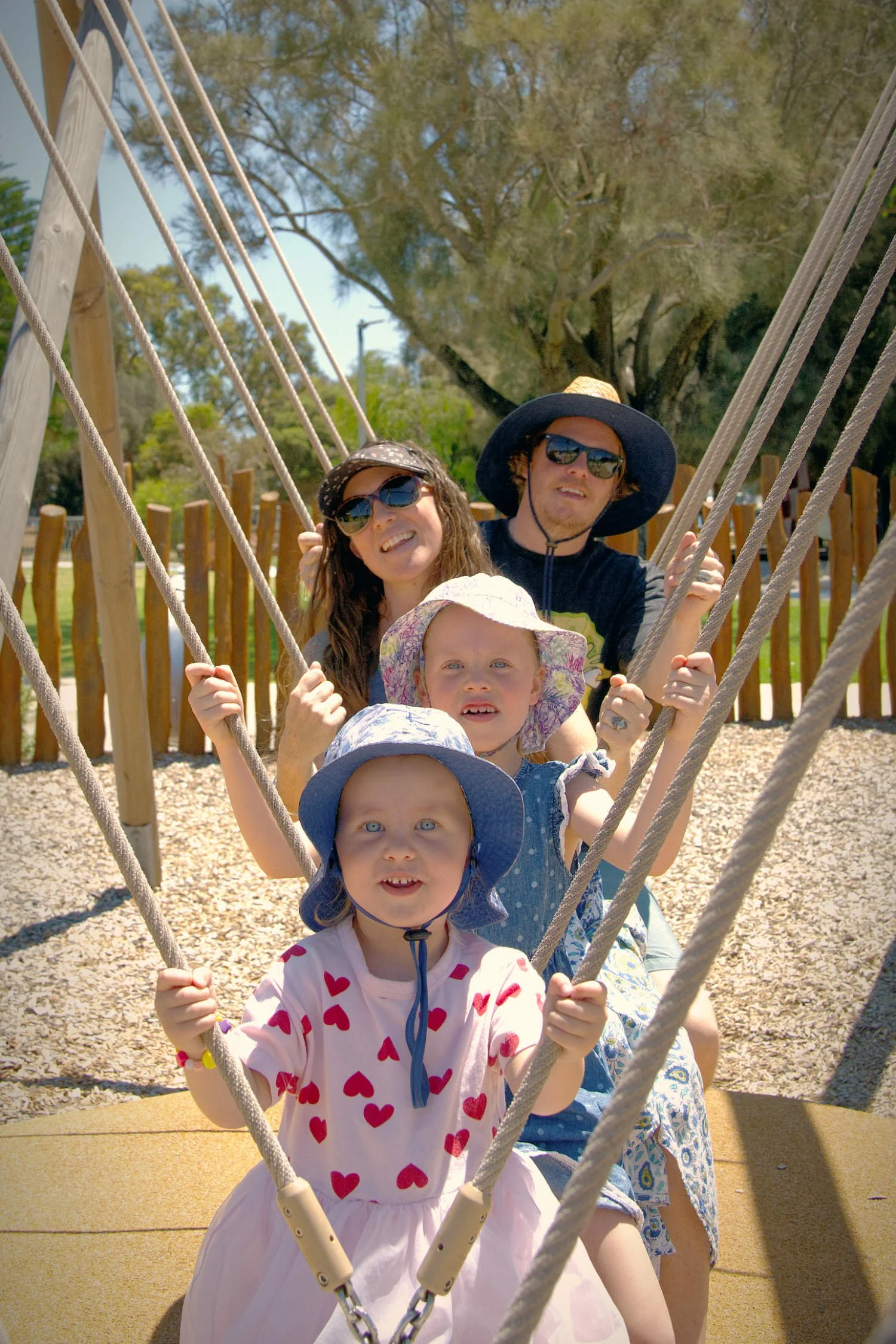 A family of four, including young children and two adults, sitting on a swing set outdoors on a sunny day. The image is in black and white.