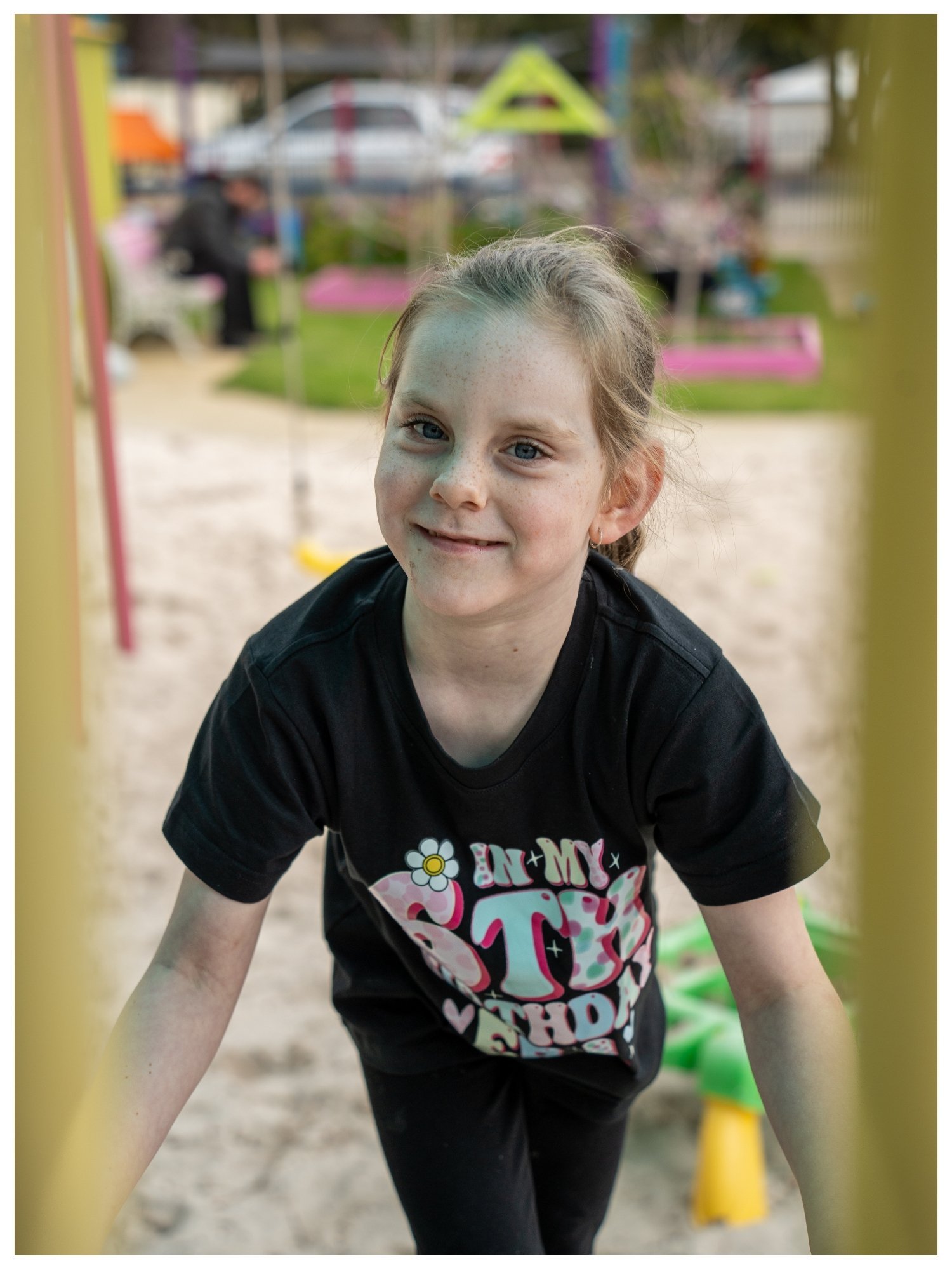 Young girl smiling at the camera, leaning forward at a playground, with playground equipment in the background.