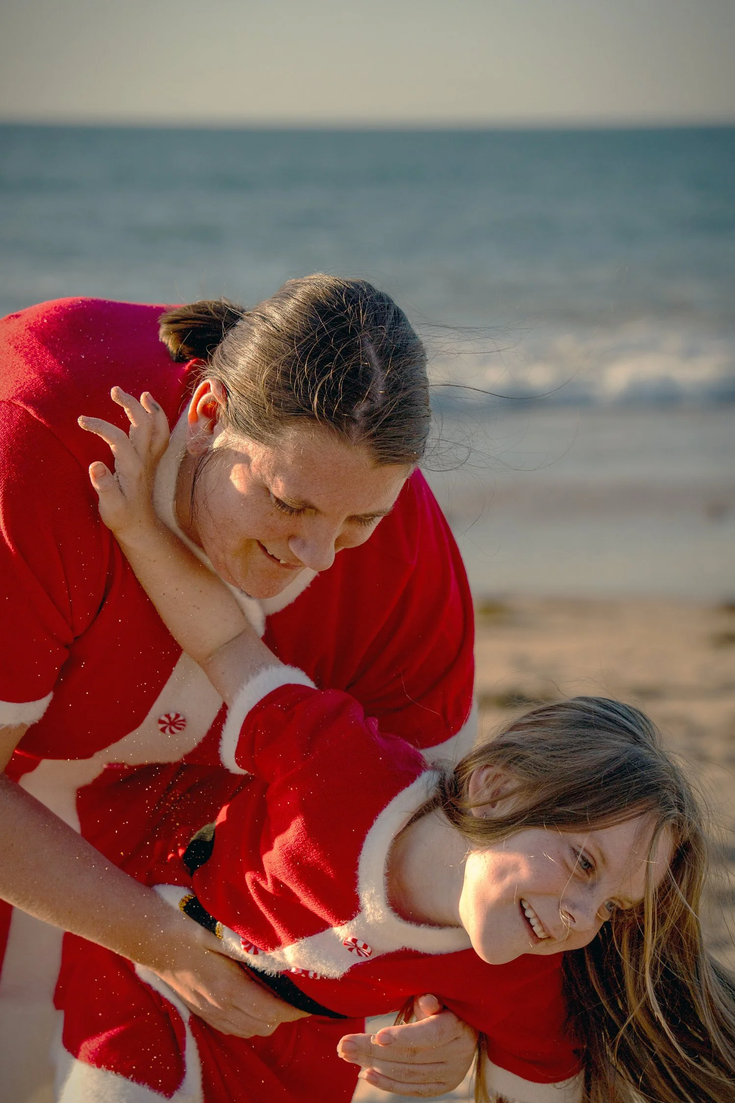 A girl and her mother play and laugh together at the beach. Both wear red Christmas dresses