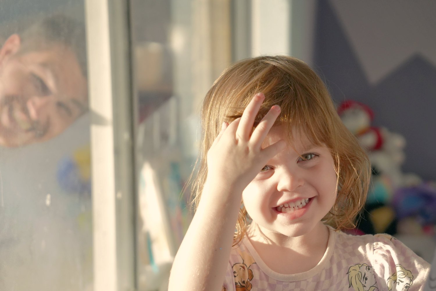 A girl twirls her hair around her finger and smiles at the camera. Behind her, her father grins through a window