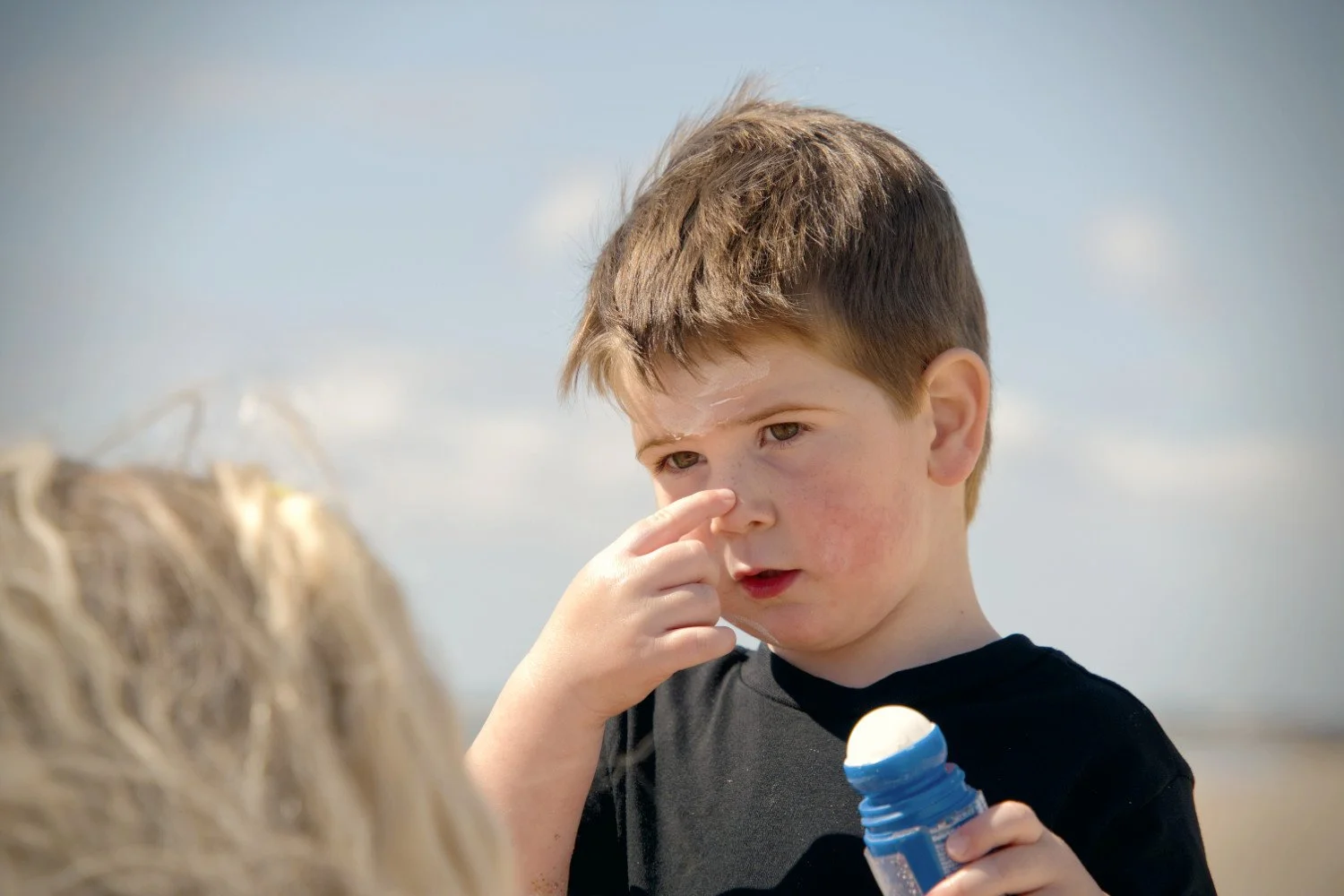 A boy holds sunscreen and points at his nose whilst looking at a blonde haired woman in the foreground
