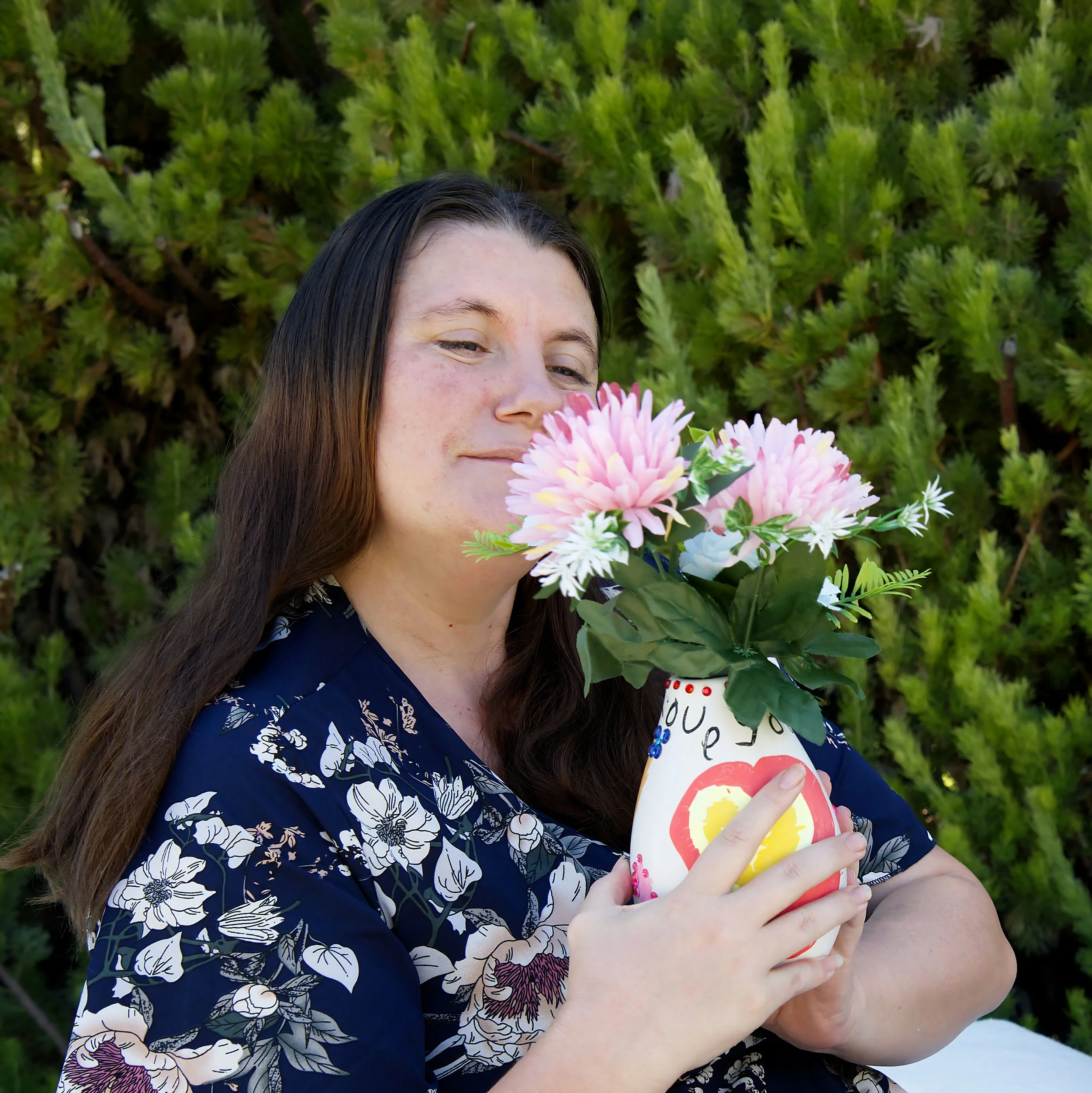 A photograph of a woman with brown hair in a dark blue dress with white and pink florals smiles as she looks at a vase of flowers decorated by her child