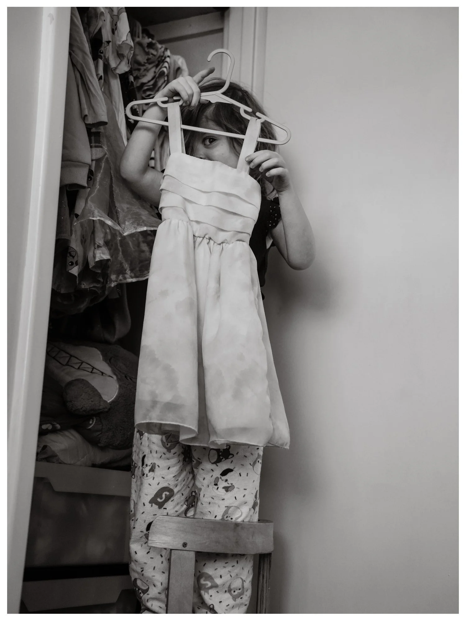 A young girl standing on a chair, holding up a dress on a hanger in front of her face, peeking over it and looking at the camera. She is in a closet with various clothes hanging on the wall beside her.