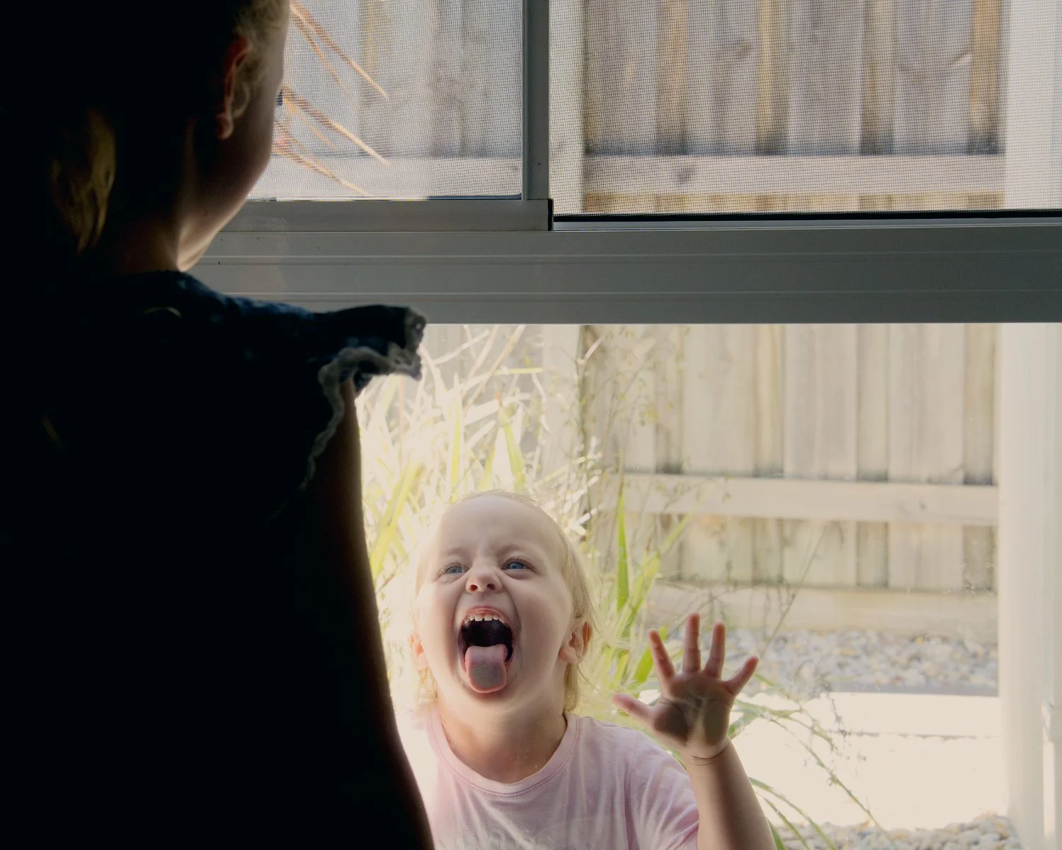 A young girl presses her face and hand against a glass door, making a silly face with her tongue out and her eyes wide open, while looking outward. 