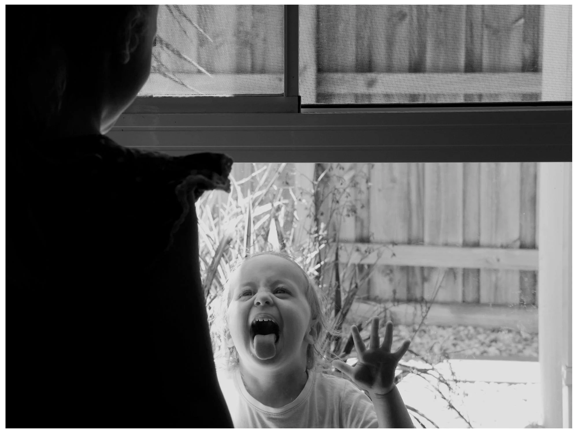 A young girl presses her face and hand against a glass door, making a silly face with her tongue out and her eyes wide open, while looking outward. 