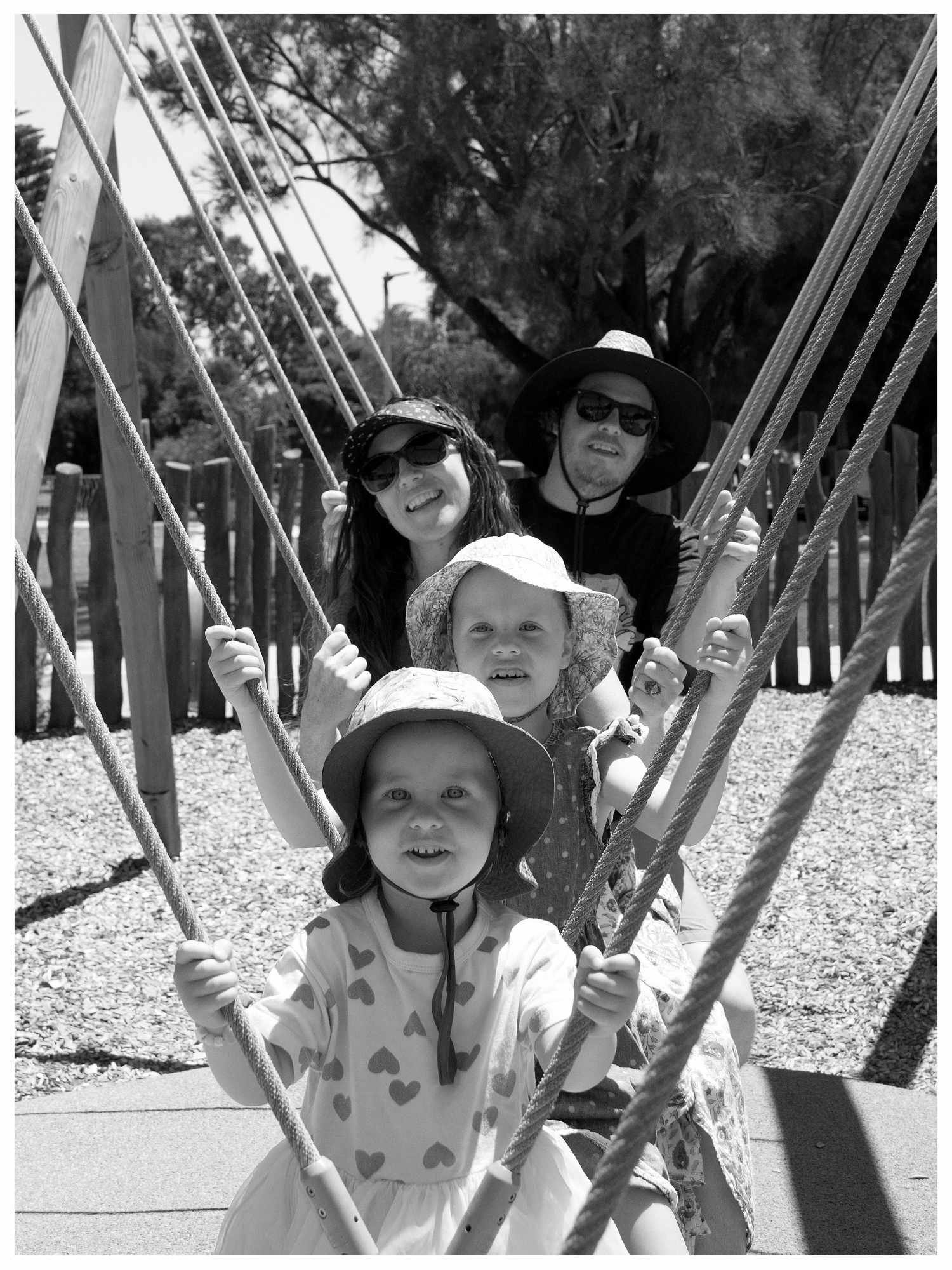 A family of four, including young children and two adults, sitting on a swing set outdoors on a sunny day. The image is in black and white.