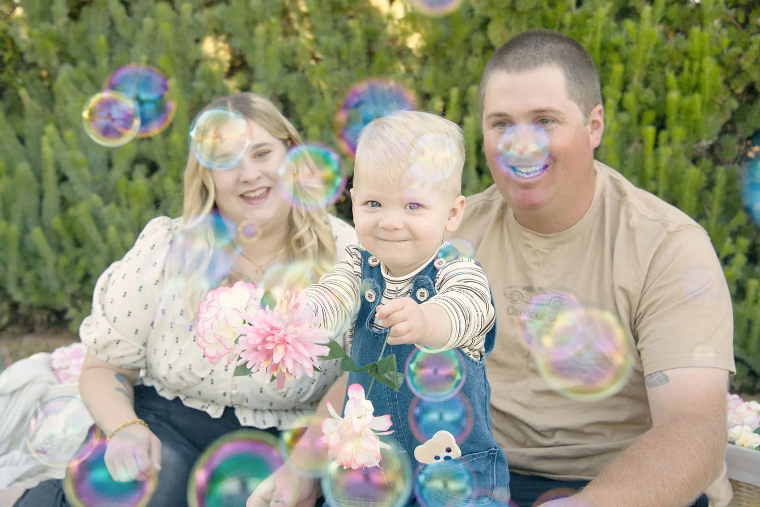 A young blonde boy in denim overalls smiles surrounded by bubbles. In the background, his parents watch and smile