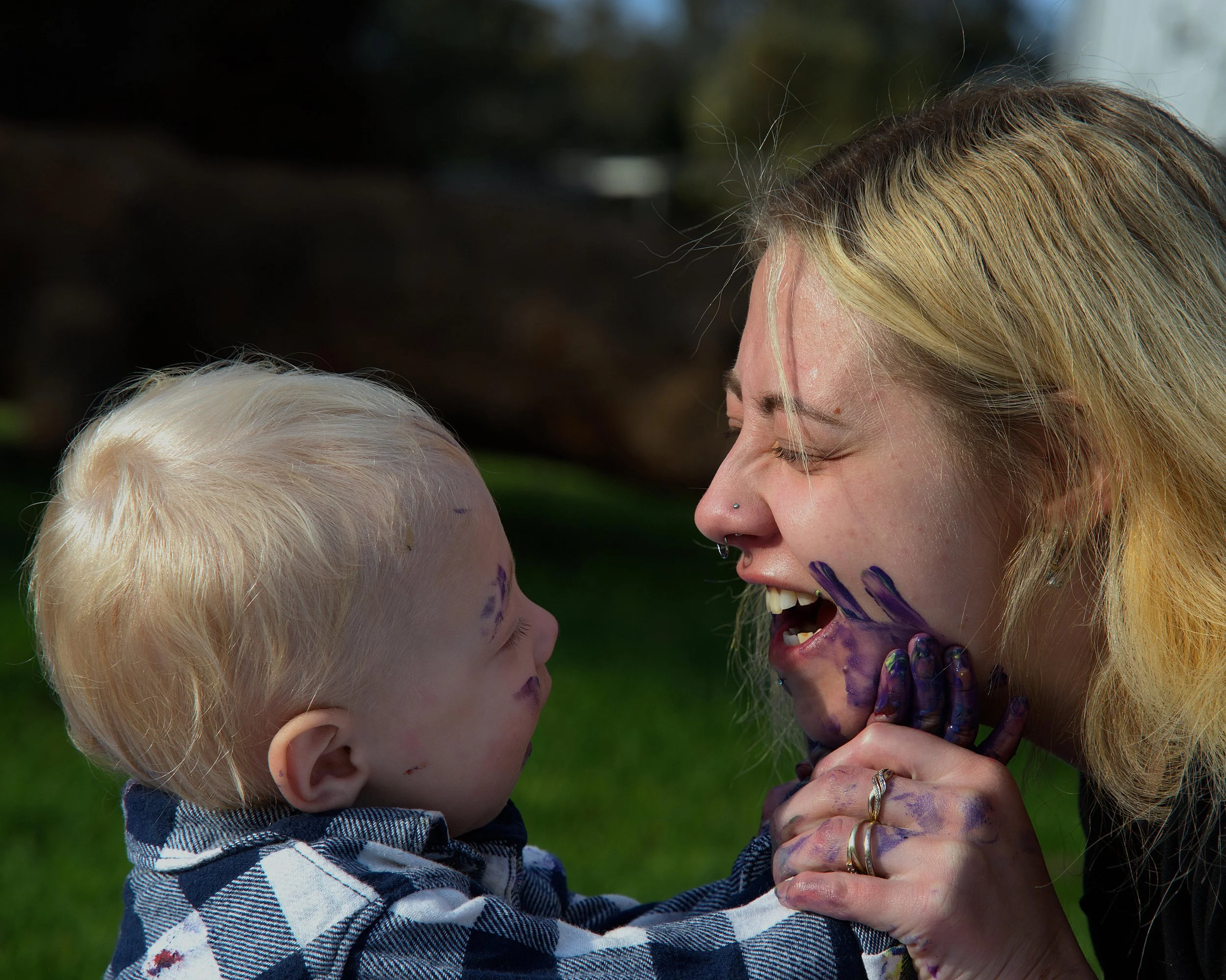 A woman and a young child playfully covered in purple paint, smiling and enjoying each other's company outside.