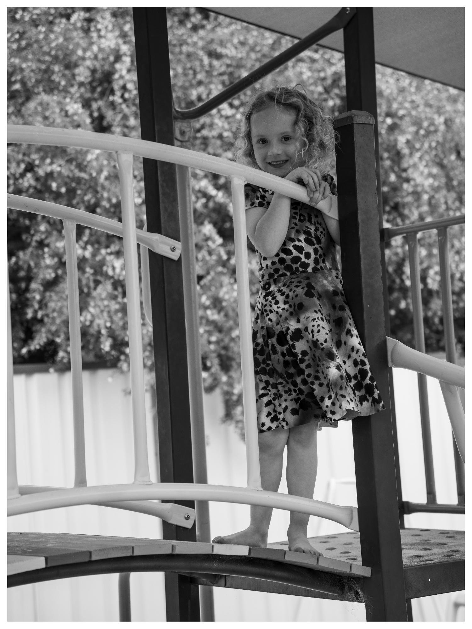 A young girl with curly hair on a playground, holding onto the railings, wearing a leopard print dress, smiling, outdoors with trees and a fence in the background.
