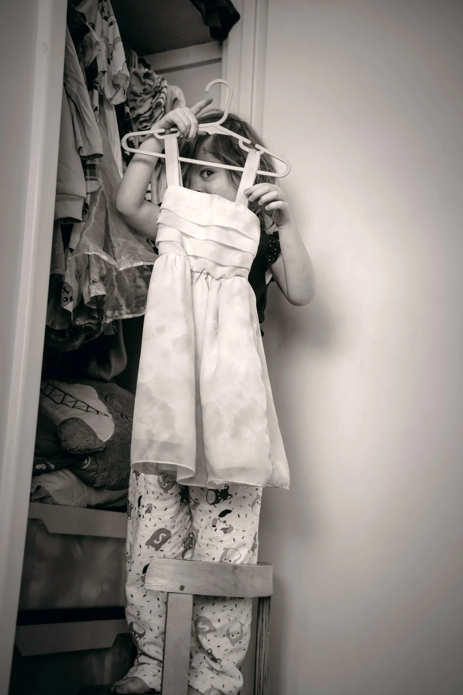 A girl in pjamas stands on a chair and holds up a dress. She peers through the hanger. a documentary family photograph in black and white by Stacey Potter