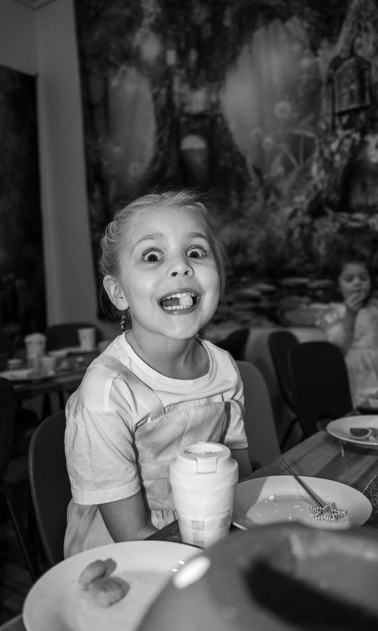 A young girl with a big smile and wide eyes making a funny face at a birthday party or gathering, sitting at a table with snacks and drinks, with another girl blurred in the background.
