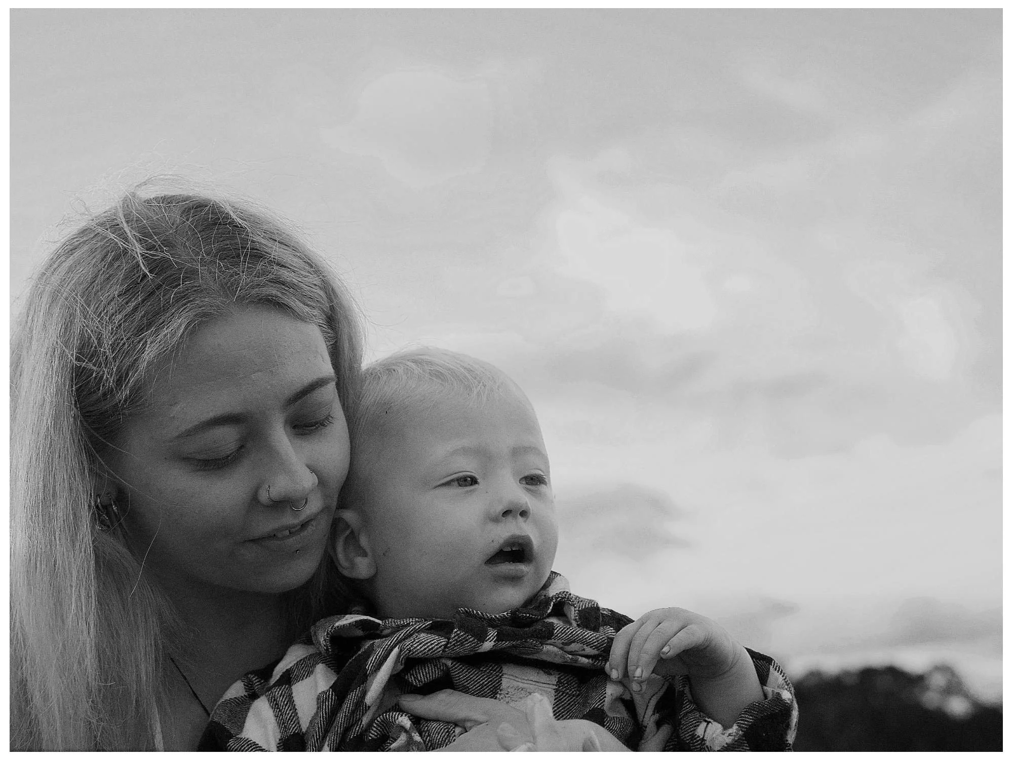 A woman holding a young child outdoors, both looking into the distance with a cloudy sky in the background.