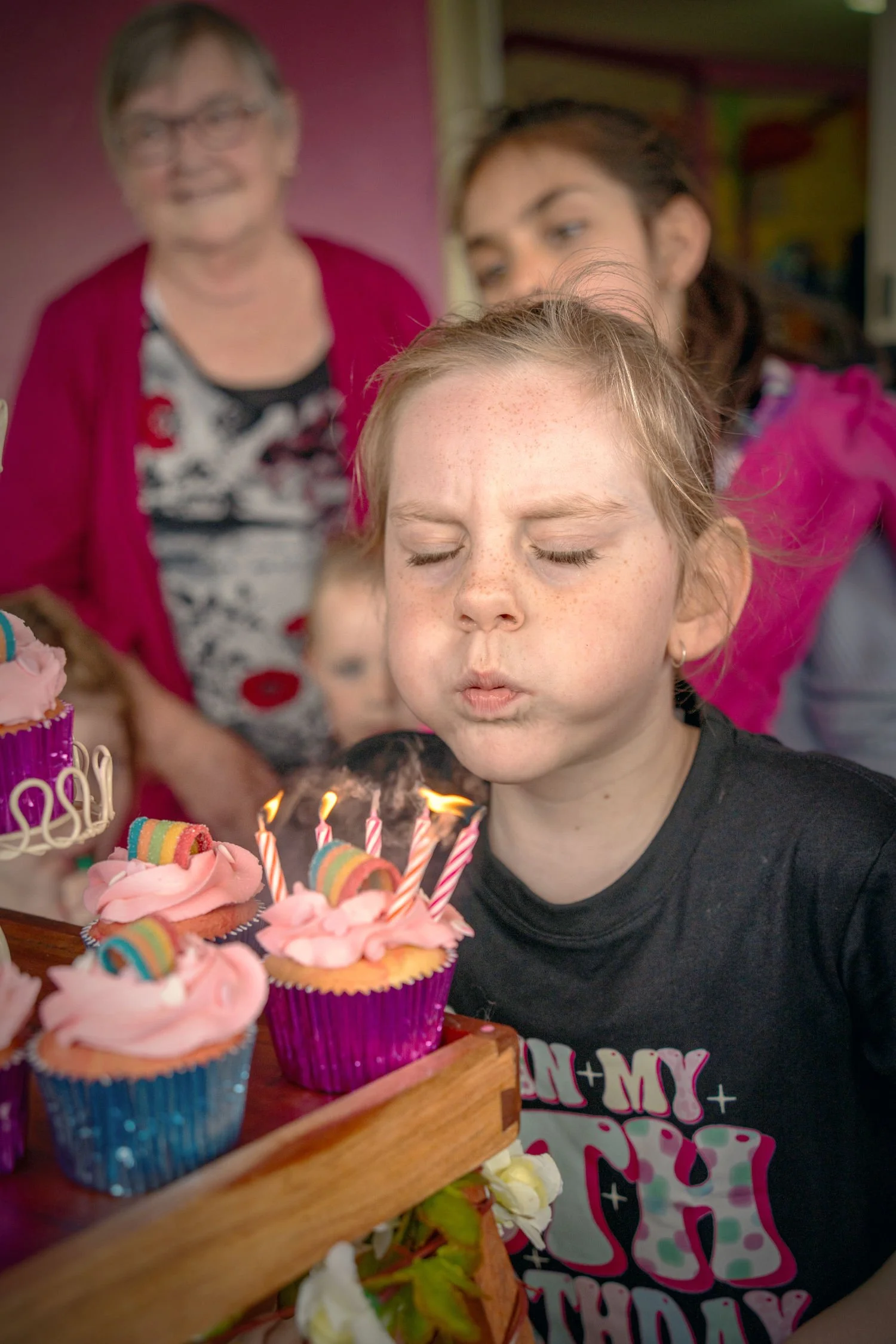 A girl with light skin, freckles, and closed eyes is blowing out candles on pink frosted cupcakes. Two women and a child are in the background, watching the celebration.