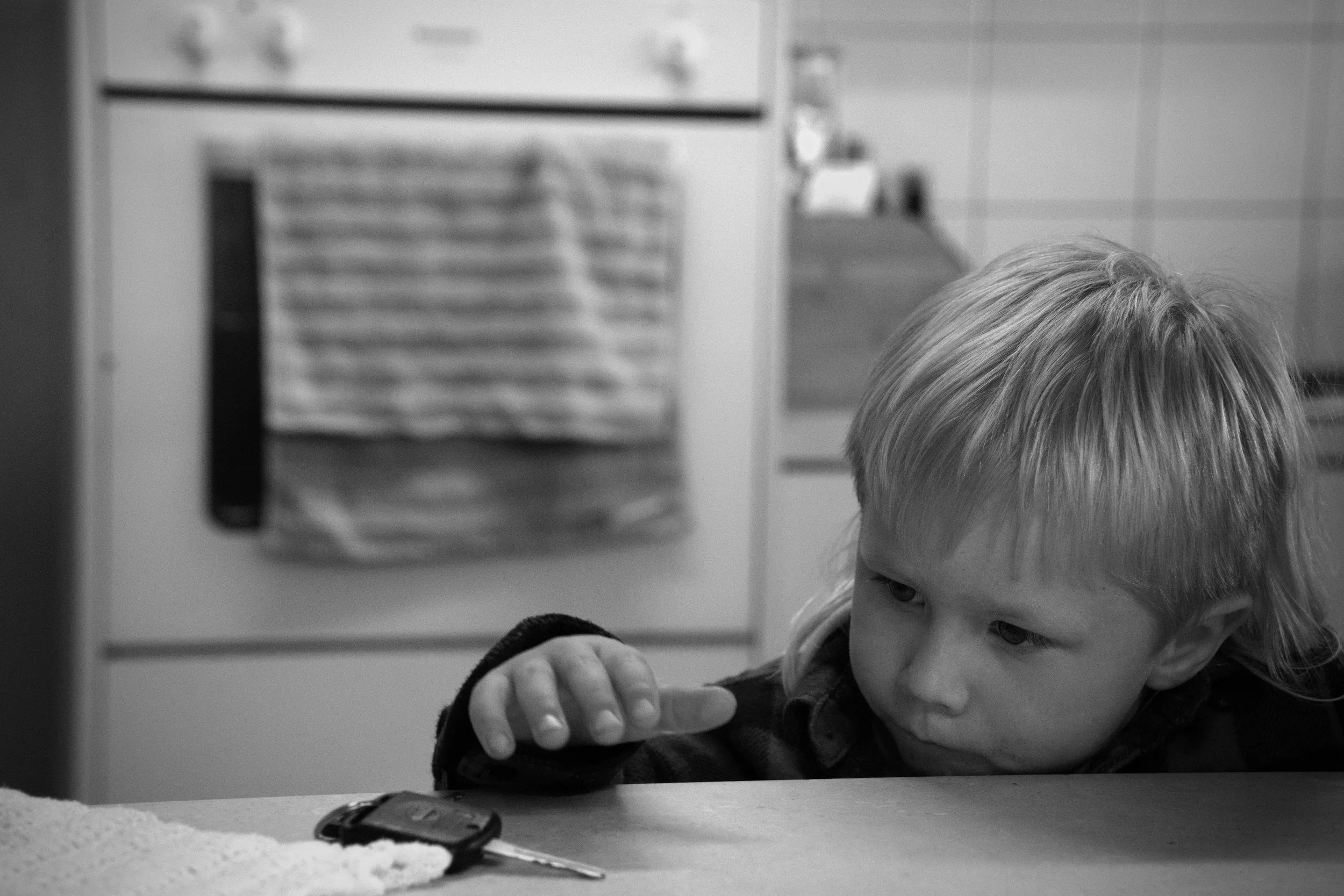 A young child with blonde hair sitting at a table, reaching out towards a set of car keys, with a kitchen in the background.