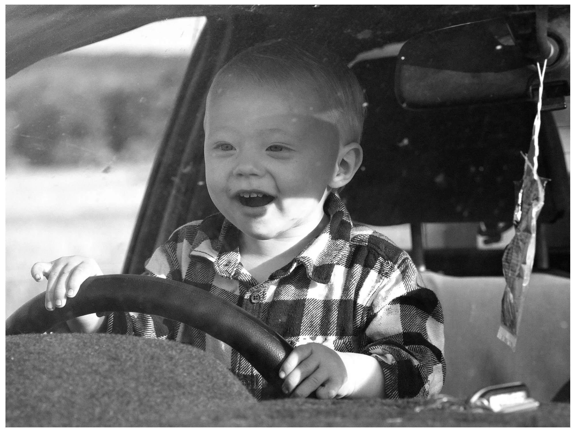 A young boy with short hair, wearing a plaid shirt, happily sitting in the driver's seat of a vehicle, holding the steering wheel, with a blurry outdoor background visible through the window.