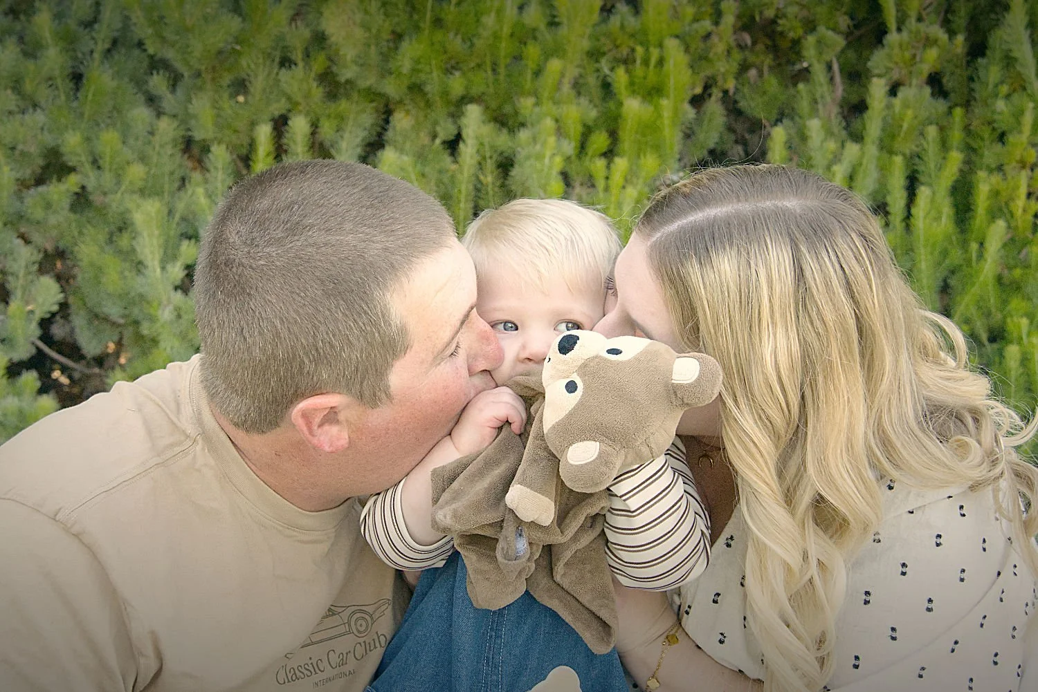 A boy holds his comforter whilst his mum and dad kiss his cheeks