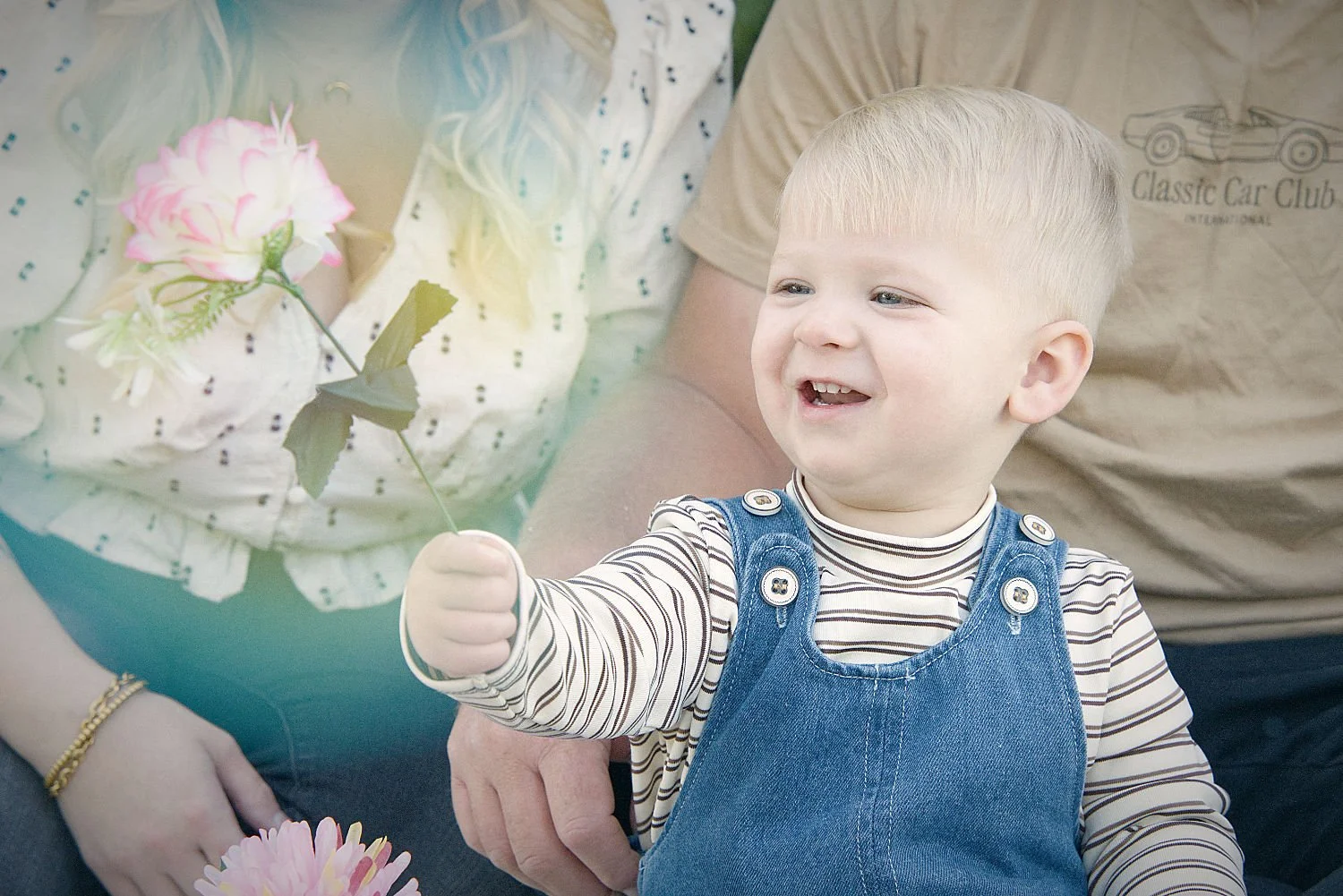 A young blonde boy  in denim overalls holds a flower and smiles