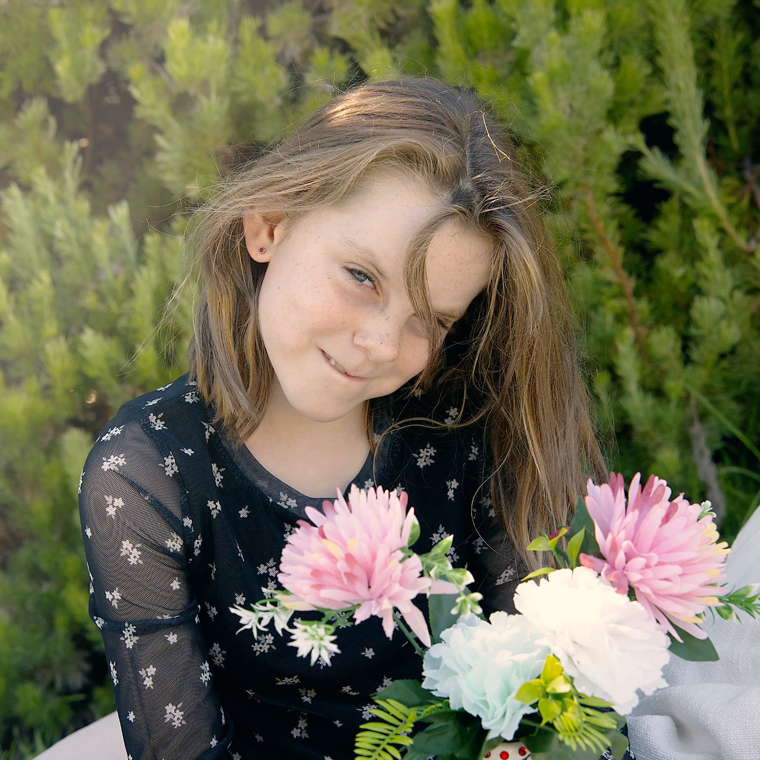 A girl with brown hair and freckles smiles shyly at the camera from behind a bunch of flowers