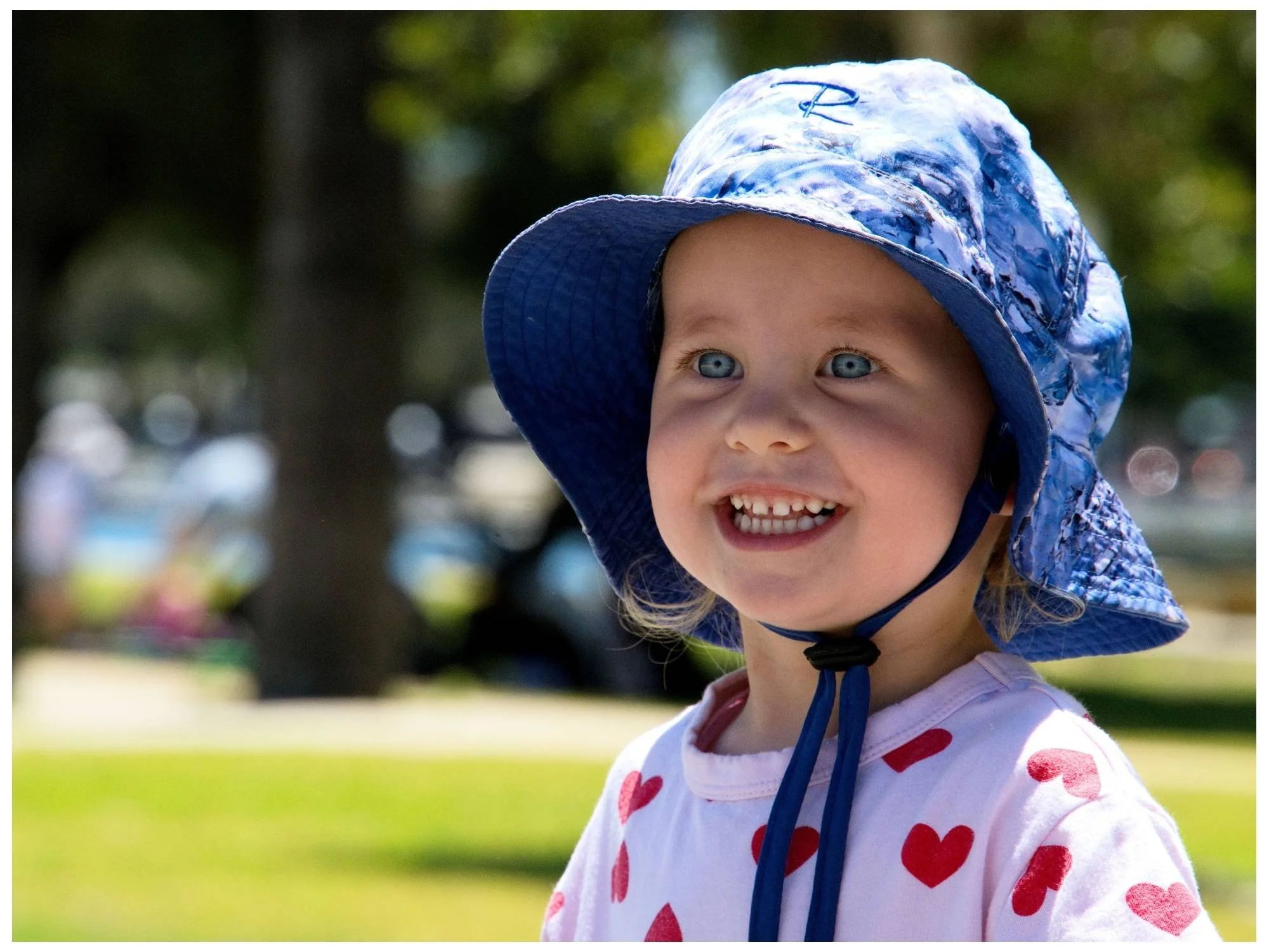 A smiling young girl with blue eyes wearing a blue and white patterned sun hat and a white shirt with red hearts, standing outdoors with trees and green grass in the background.